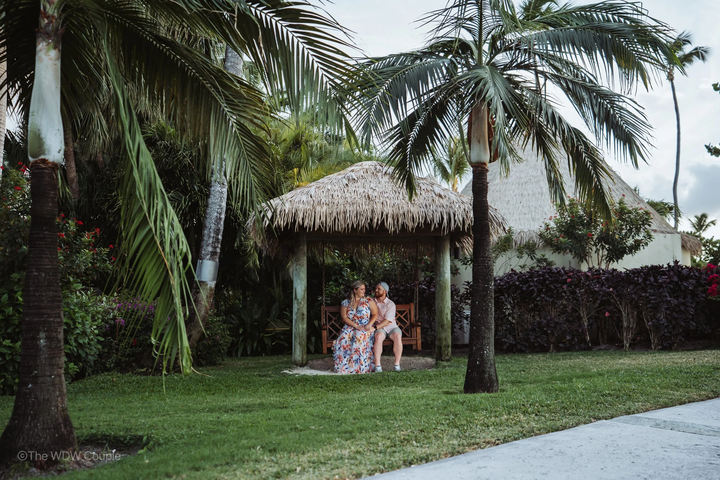 A couple sits on a wooden bench under a thatched roof gazebo in a tropical garden with palm trees and colorful plants.
