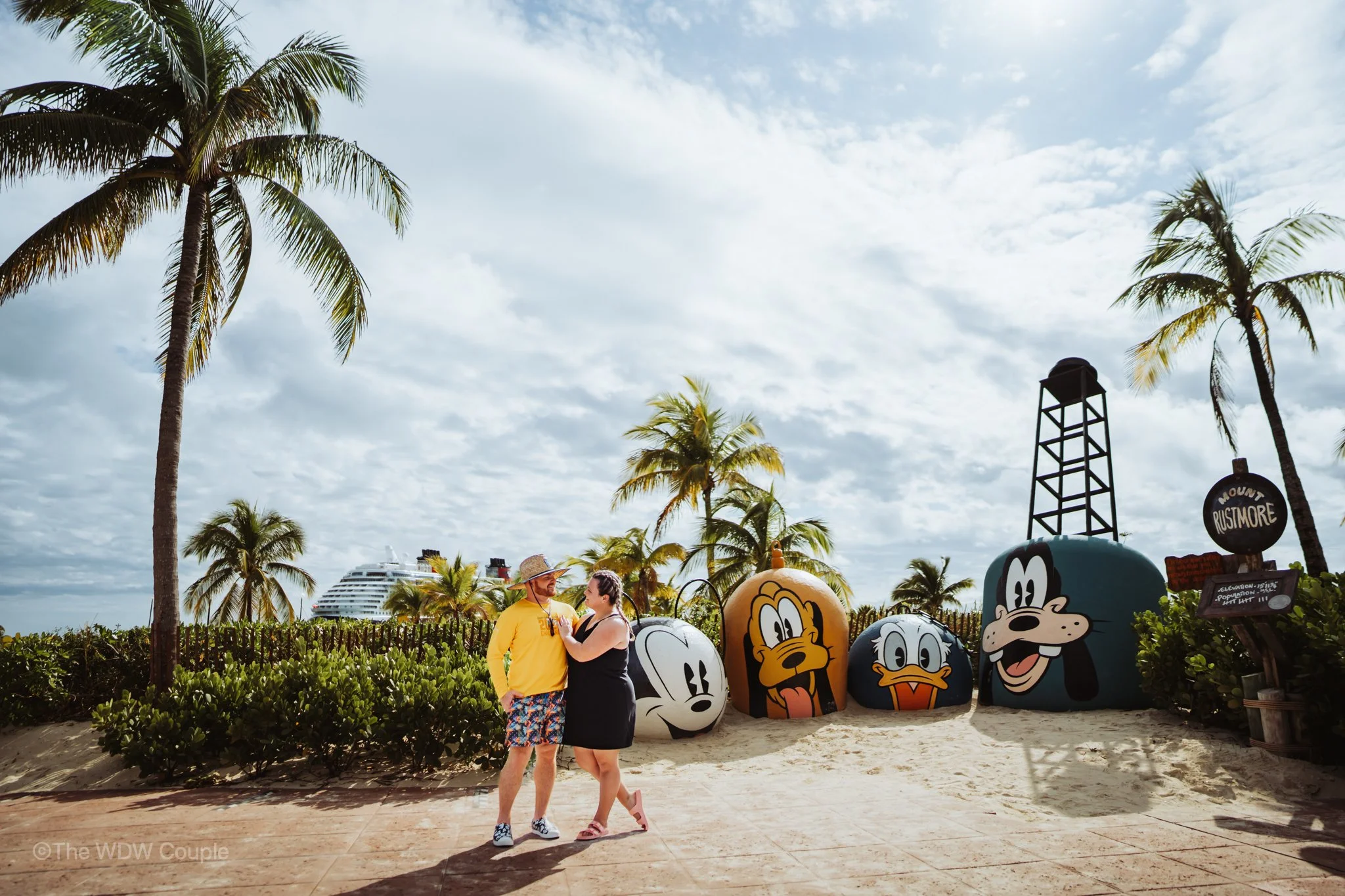A couple standing on a sandy beach pathway, looking at each other, with large cartoon character heads of Disney characters Mickey Mouse, Daisy Duck, Donald Duck, and Goofy behind them. Palm trees, a partly cloudy sky, and a cruise ship in the background.