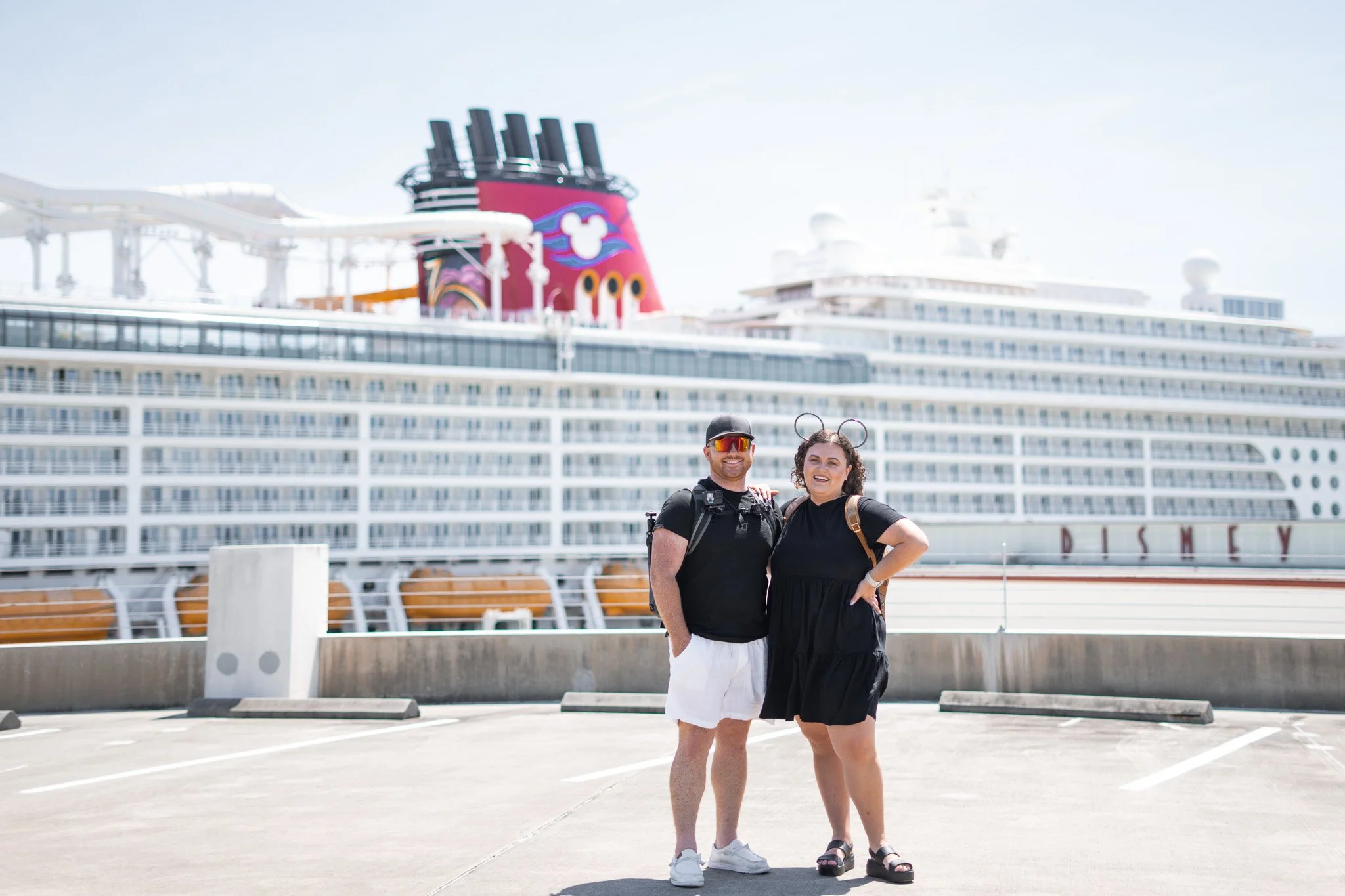 A couple stands in front of a large cruise ship at a port, with the man wearing sunglasses, a black t-shirt, and white shorts, and the woman wearing a black dress, Mickey Mouse ears, and sandals.