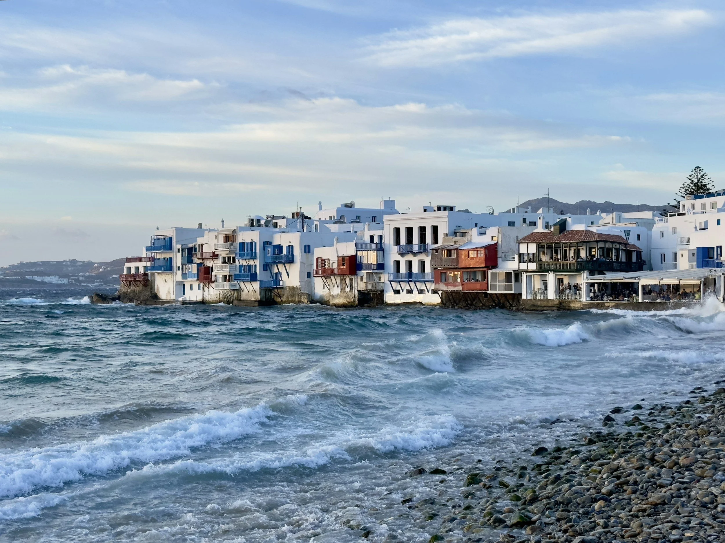 A coastal scene with white buildings featuring blue and red accents, along a rocky shoreline with waves crashing against the shore.