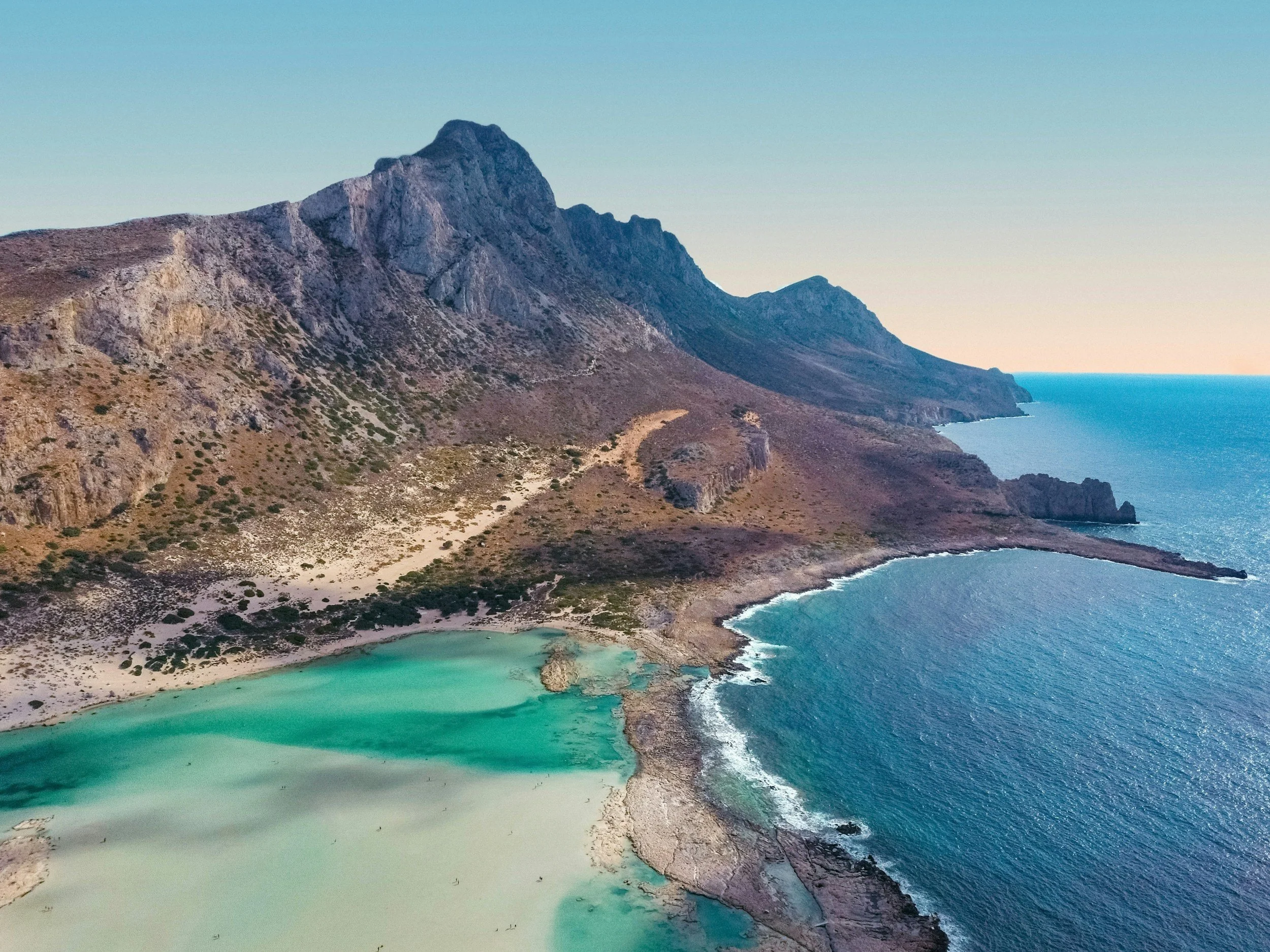 A rugged coastline with a mountain, a small inlet with turquoise water, and the open ocean under a clear sky.