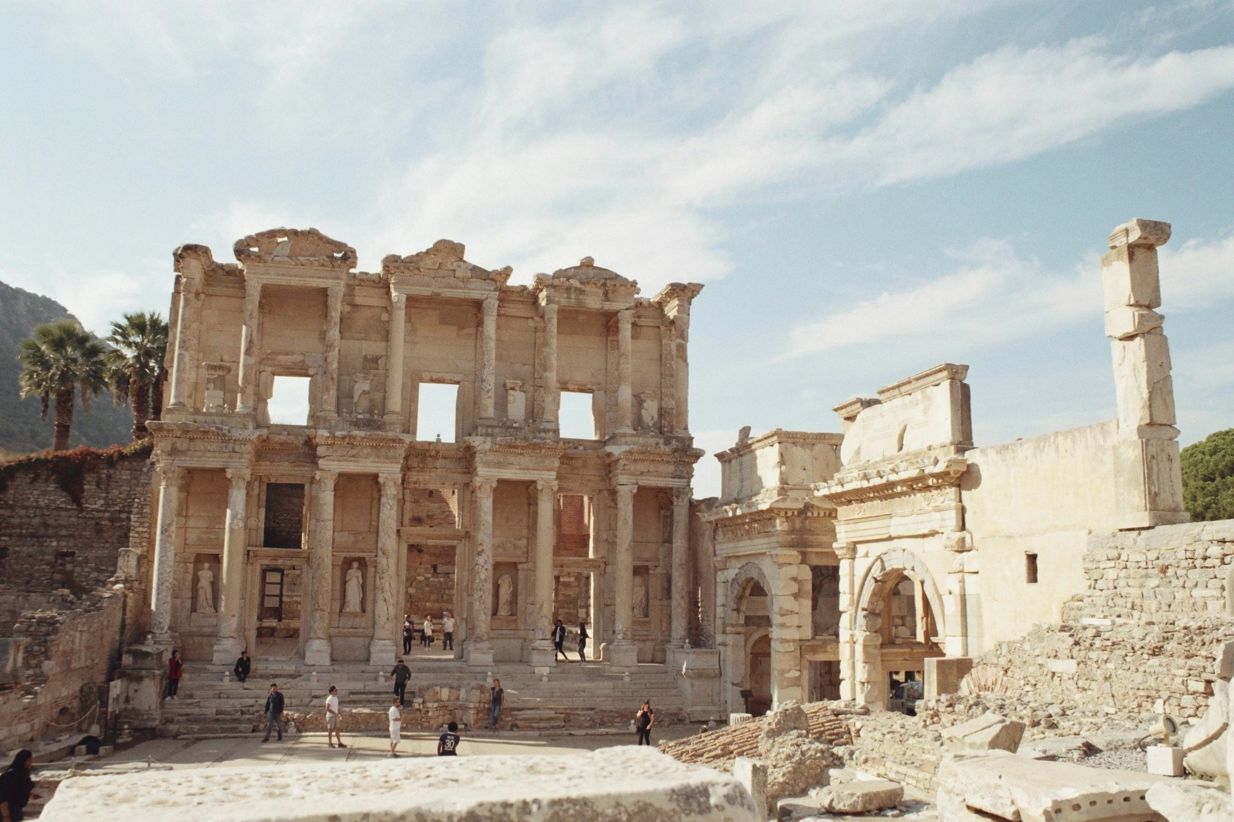 Ancient ruins of a large stone building with multiple arched windows and columns, with some parts broken or missing, under a blue sky, with a few tourists exploring the site.