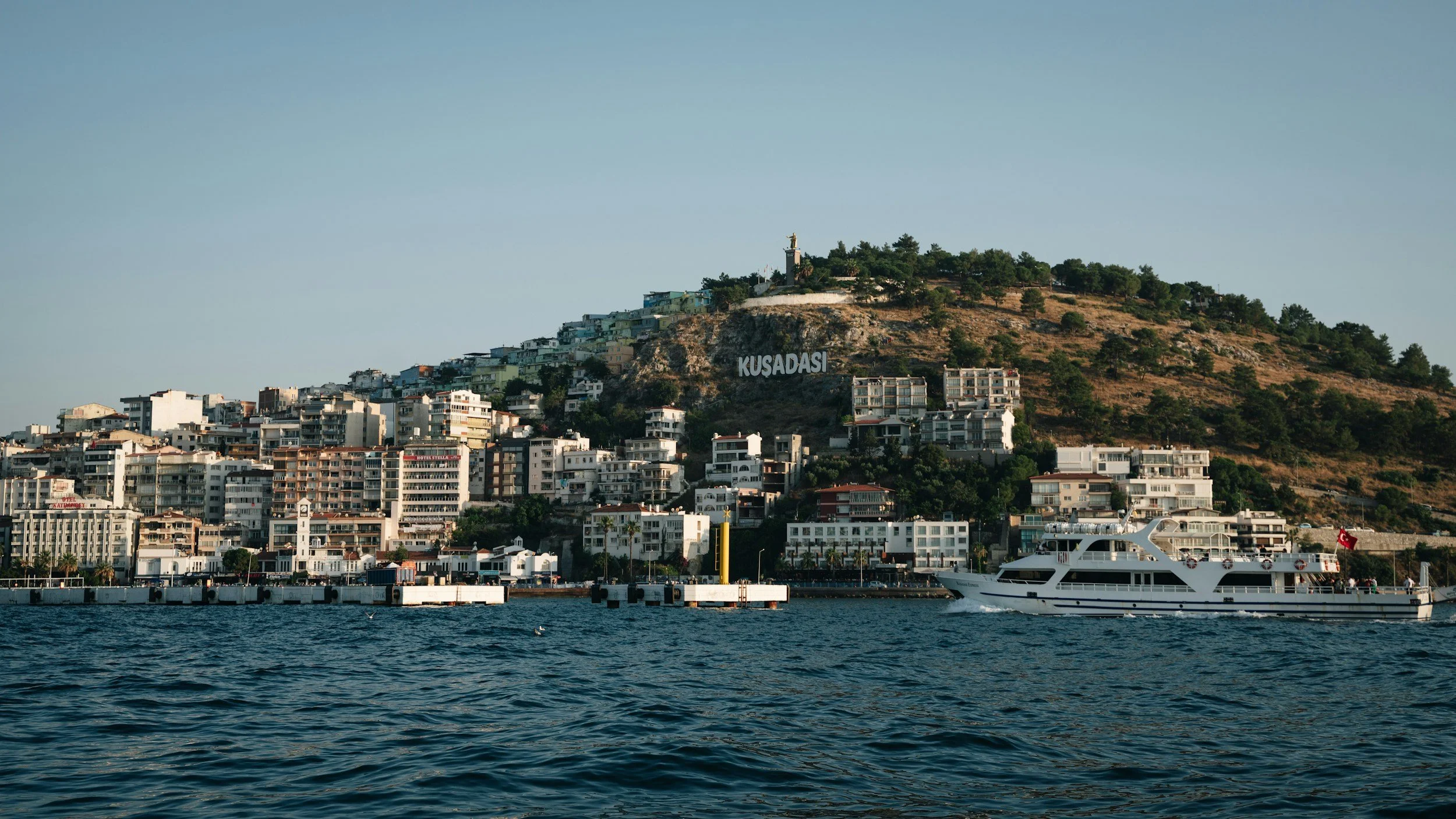 A coastal city with numerous white and beige buildings on a hillside, with the word 'Kuşadası' visible on the rocky slope. There is a large white yacht sailing on the water in the foreground.