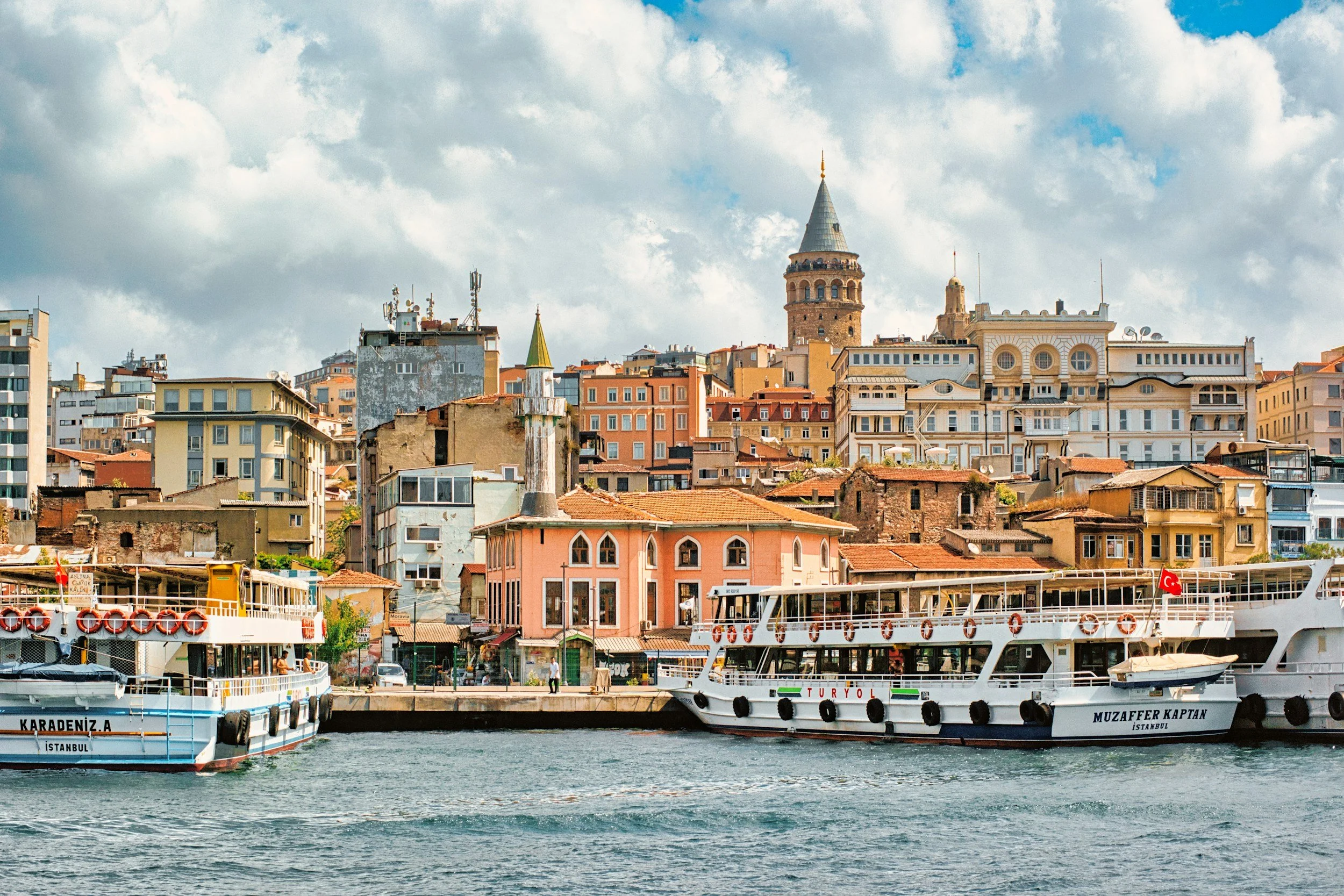 View of Istanbul skyline with historic buildings and the Galata Tower in the background, and boats docked along the water in the foreground.