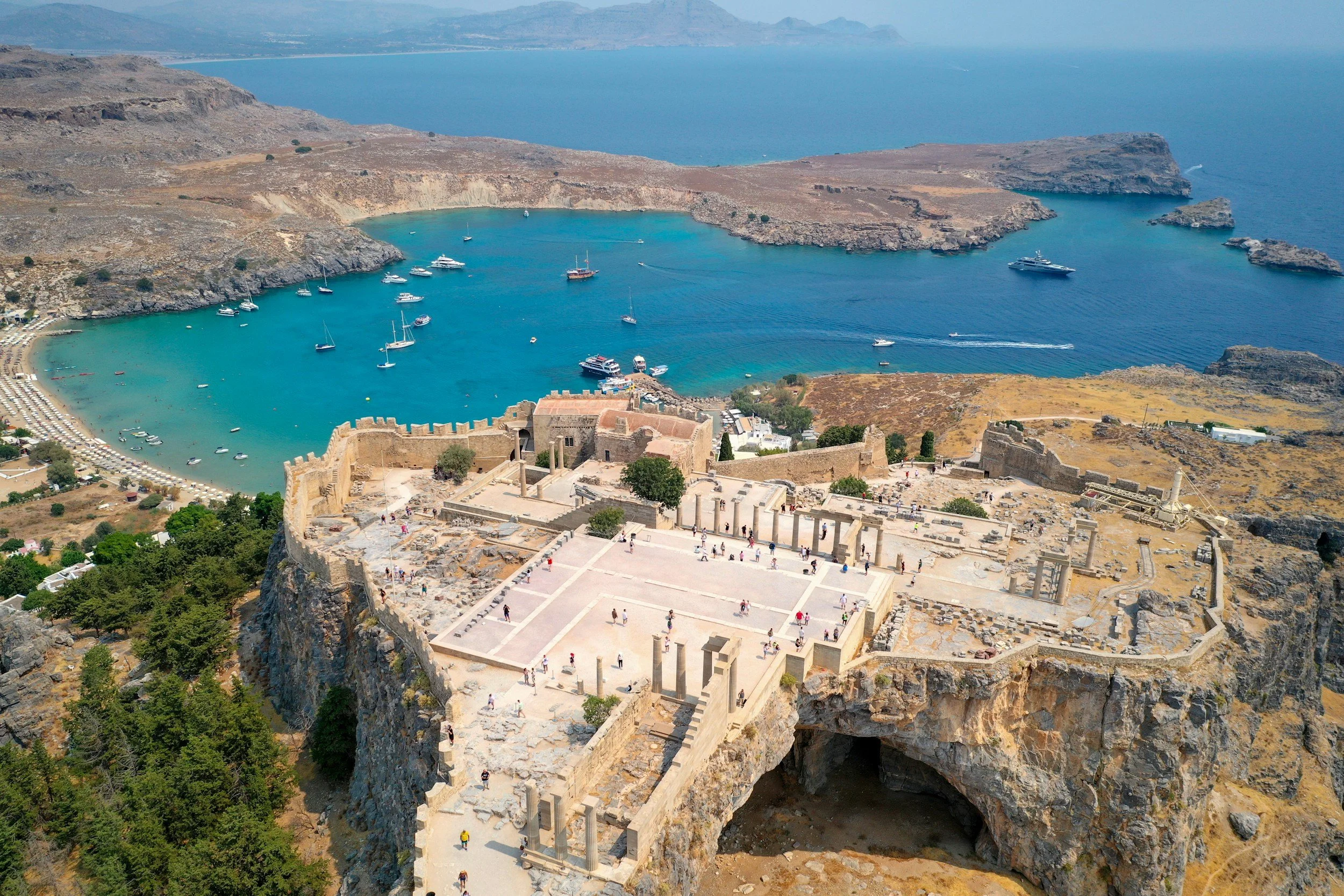 Aerial view of an ancient archaeological site with ruins and columns overlooking a harbor with boats and sailboats, along a rocky coastline with a sandy beach and a bay with turquoise water.