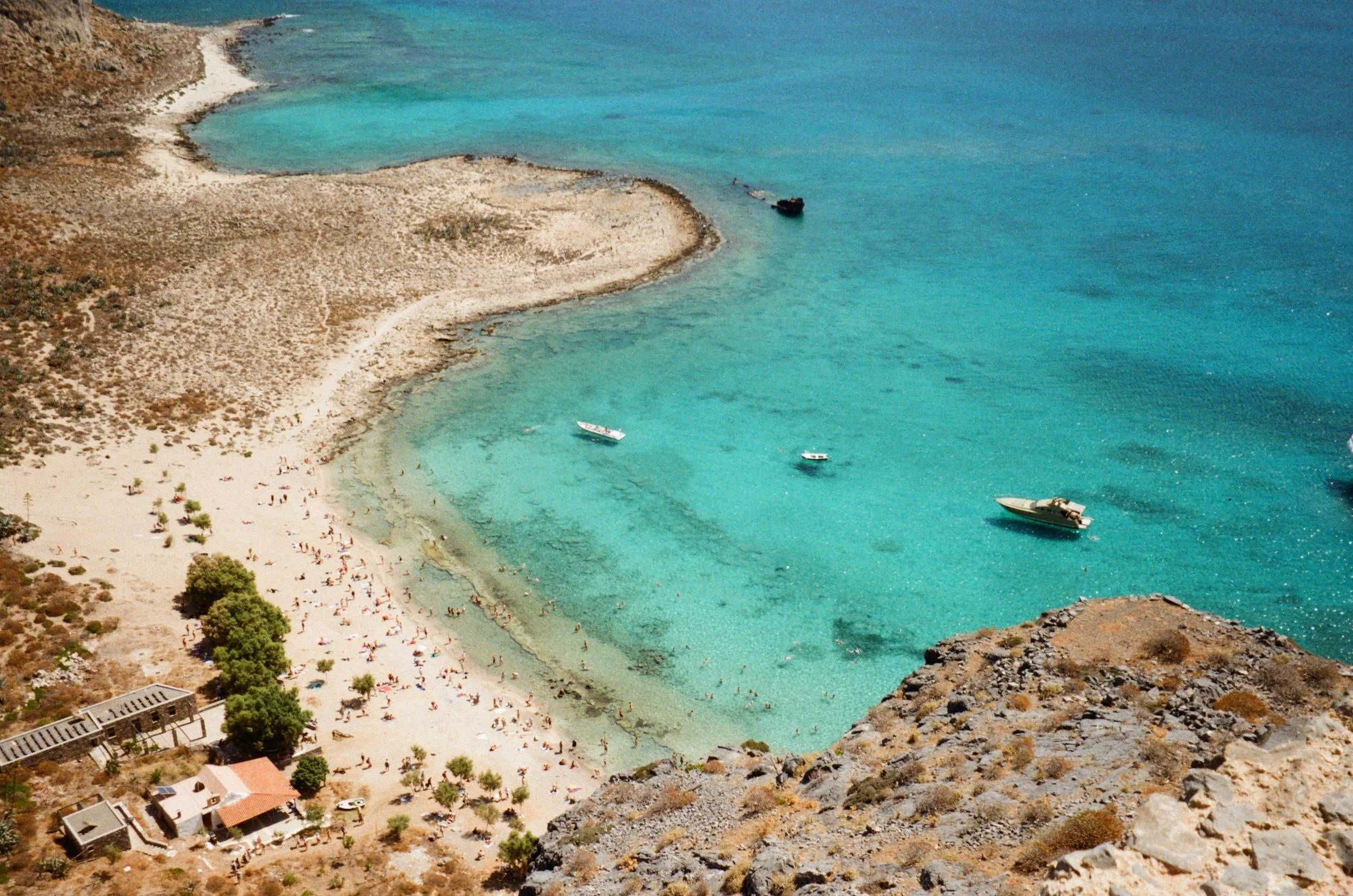 Aerial view of a beach with clear turquoise water, surrounded by rocky terrain, small boats anchored in the water, and people sunbathing and swimming on the sandy shore.