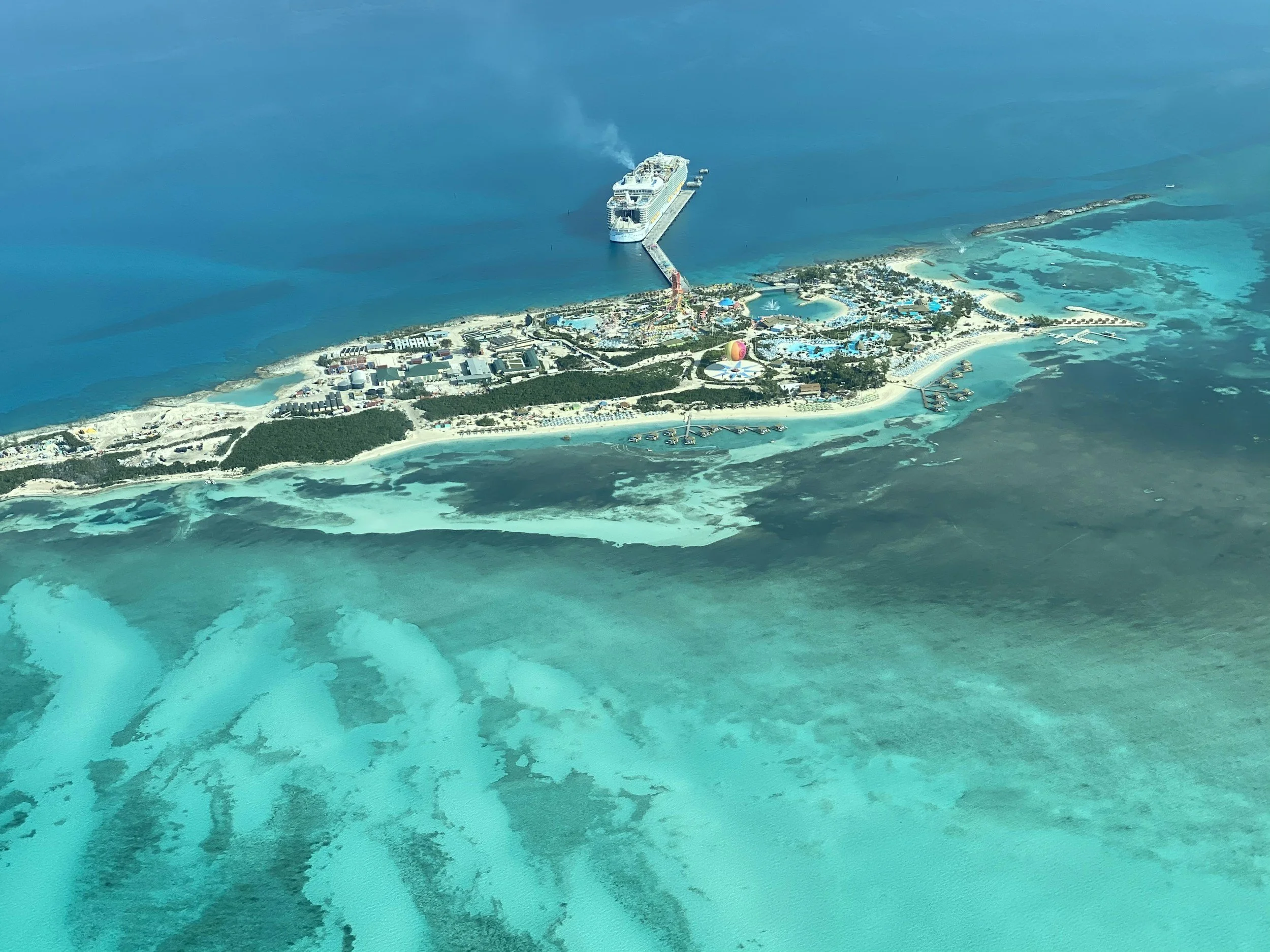 Aerial view of a tropical island with turquoise waters, a large cruise ship docked at a pier, and various buildings and attractions on the island.