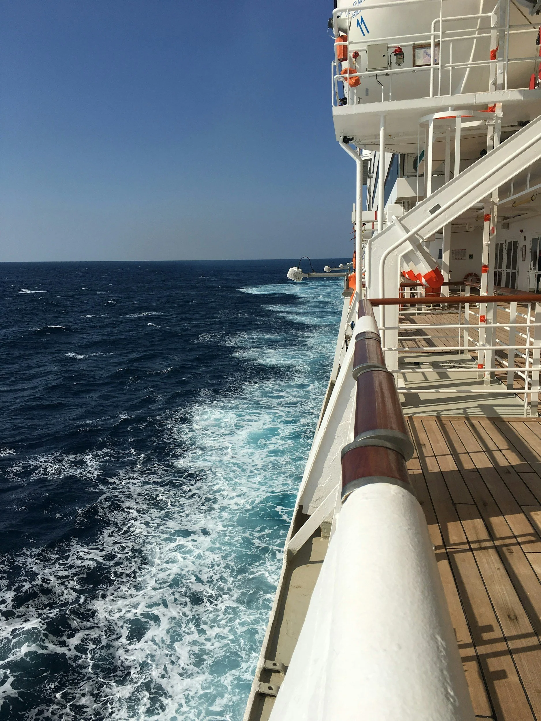 View of the ocean from the deck of a cruise ship, with white waves behind the ship as it moves through deep blue water, under a clear sky.