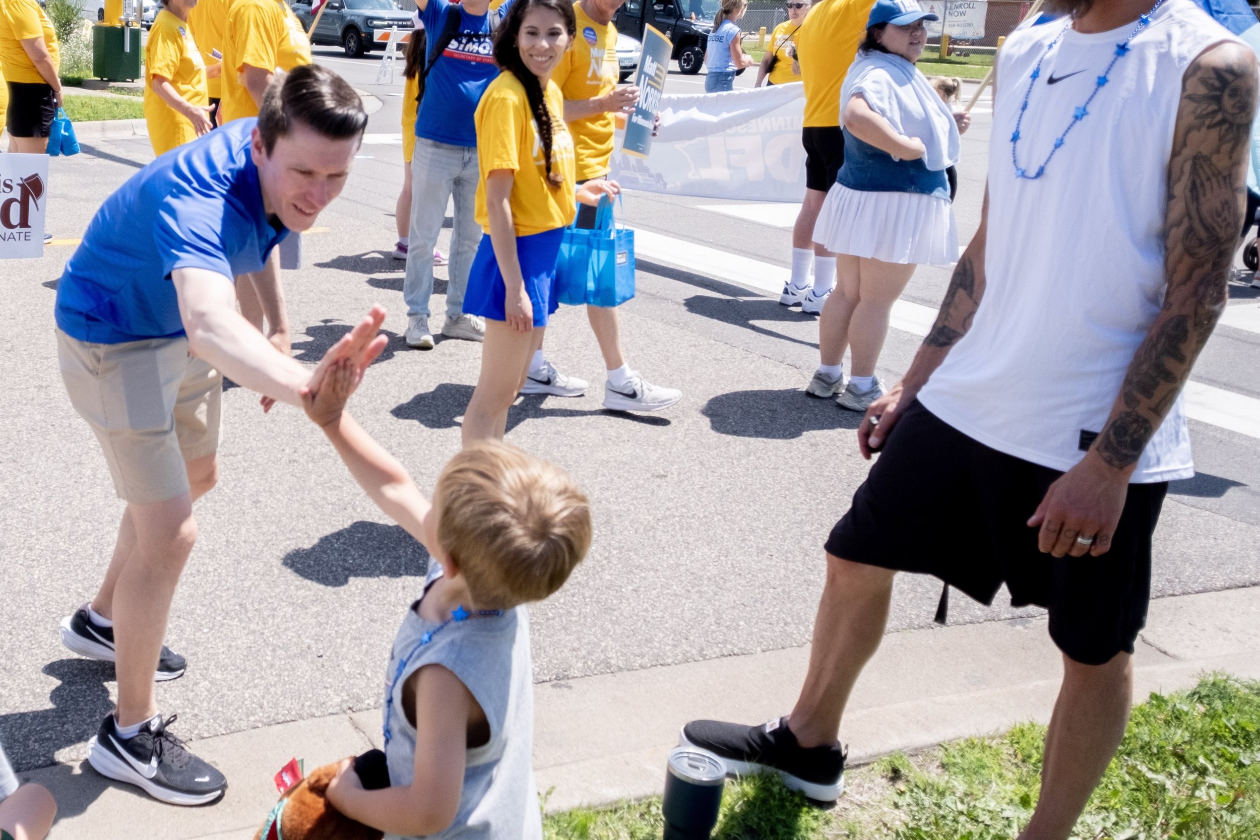 Matt Norris participating in a parade event on a sunny day, with some wearing yellow and blue shirts, some holding signs, and children interacting with adults near the sidewalk.