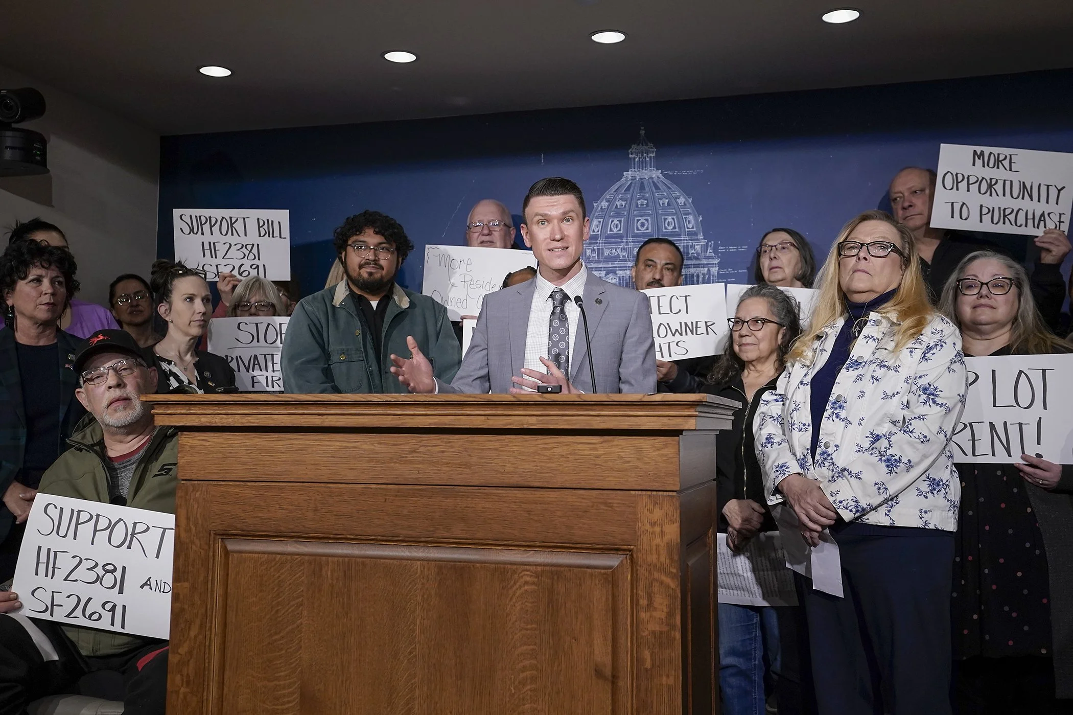 A man in a gray suit and tie speaking at a podium in front of a crowd of people holding protest signs, with a blue backdrop featuring a white outline of the United States Capitol.