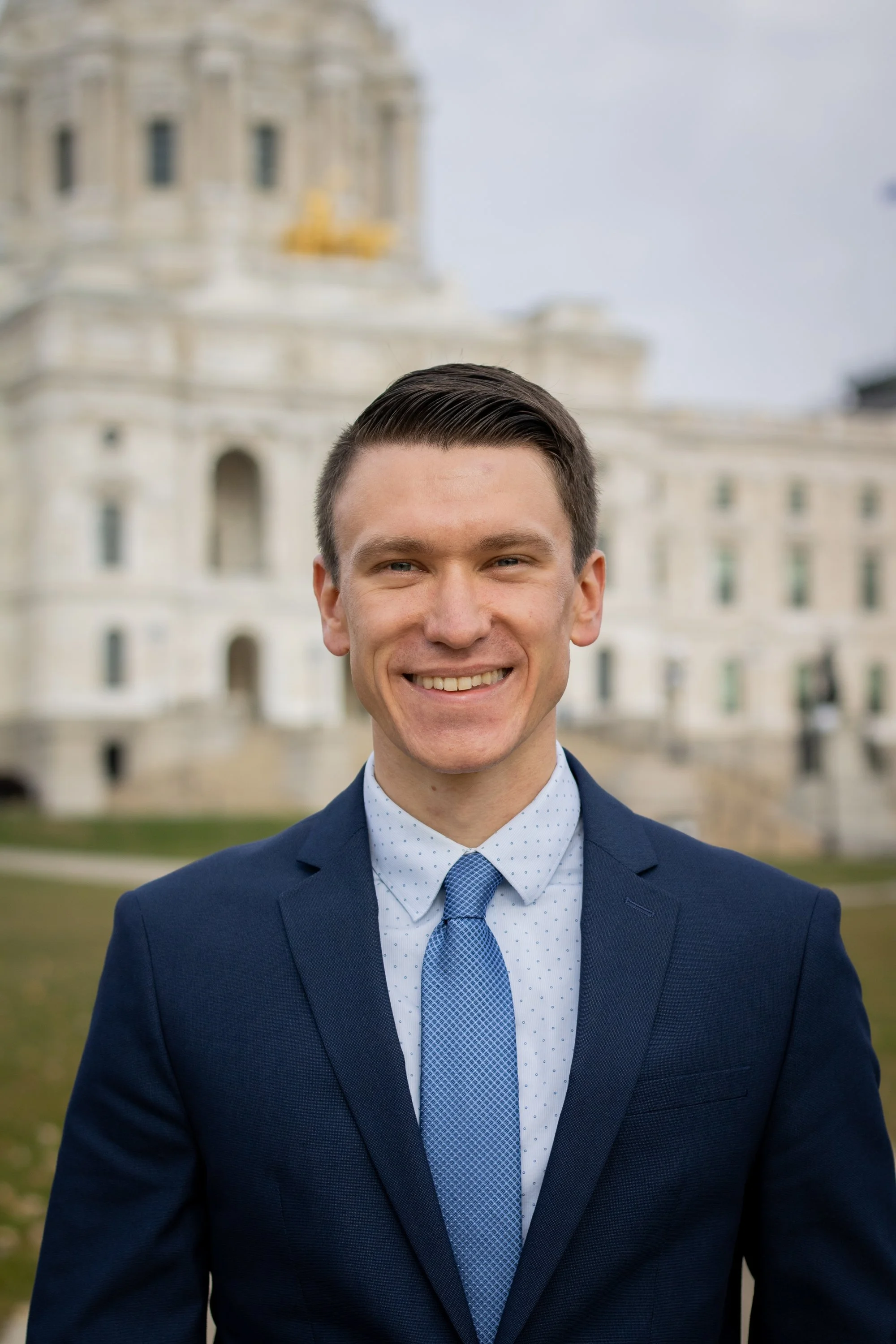 Matt Norris in a navy blue suit and light blue tie standing outdoors with the Minnesota State Capitol building in the background.