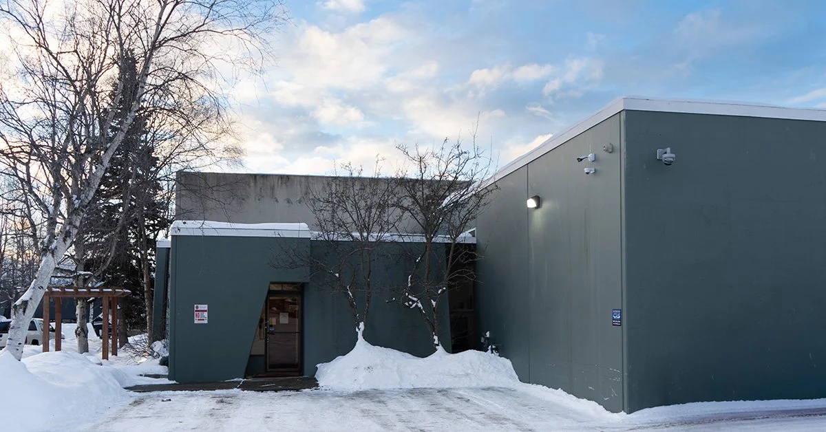 WEKA Tactical in Anchorage Firearms Shop. A modern, gray building in a snowy landscape with leafless trees, a wooden fence, and a partly cloudy sky.
