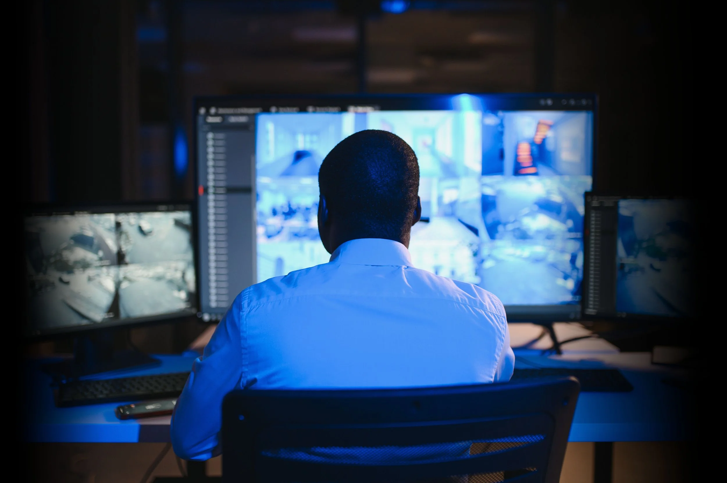 A person wearing a white shirt working at a computer with multiple monitors in a dark room, illuminated by blue light.