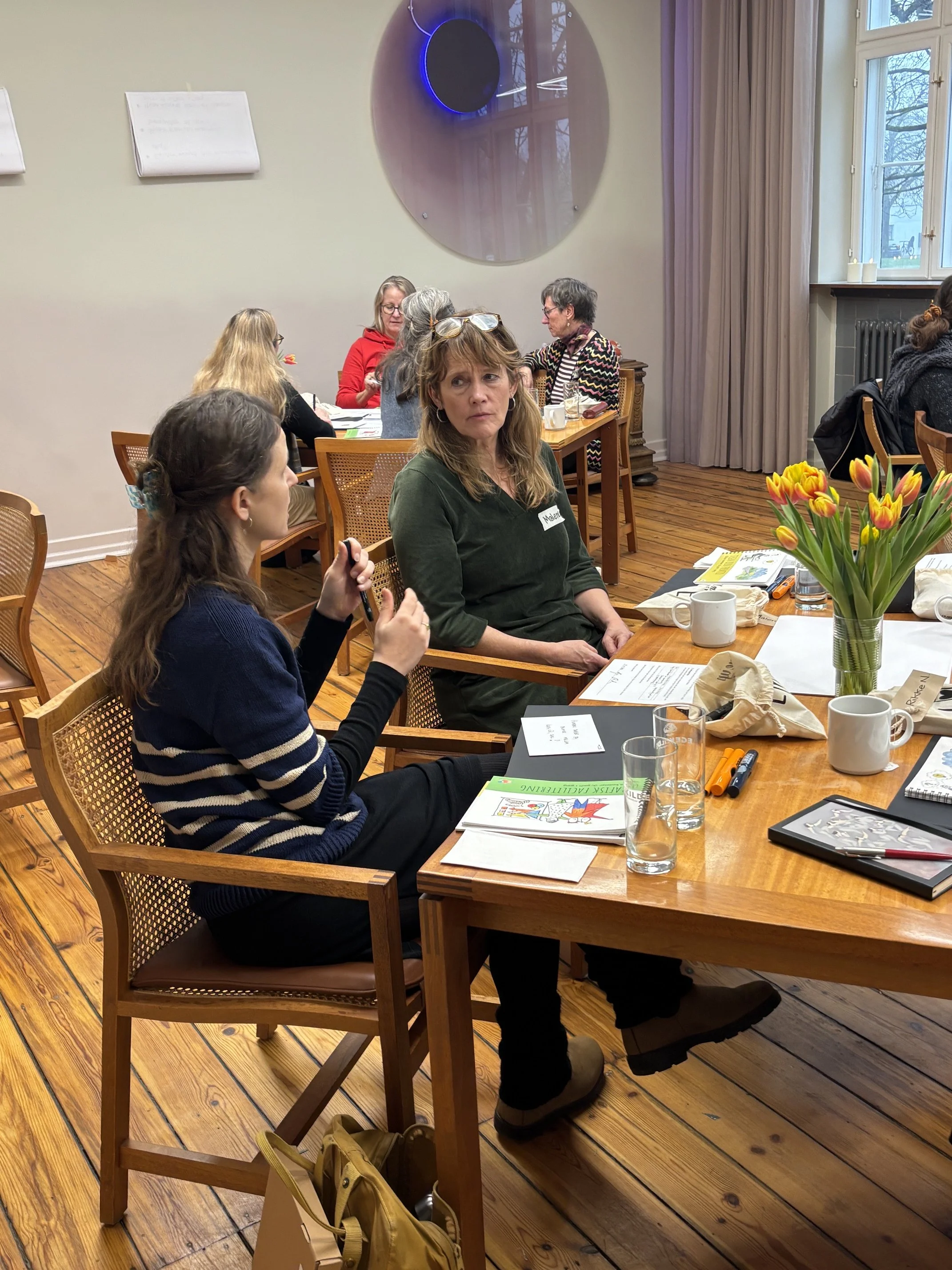 Et workshop lokale, hvor deltagerne sidder ved mindre borde og taler og lytter til hinanden
