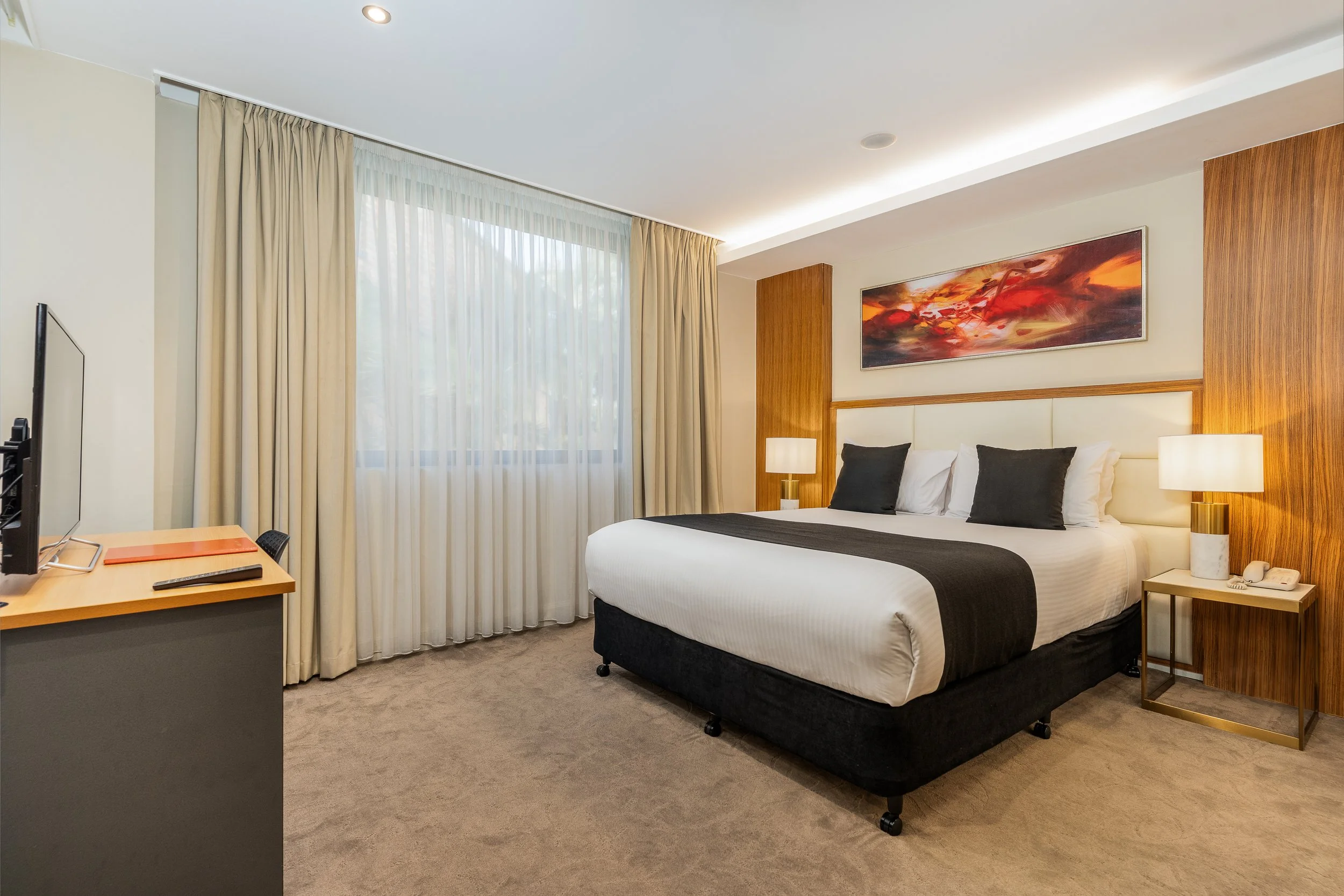 A modern hotel room with a large bed, white bedding, black and white pillows, and wooden accents. There are two lamps on nightstands, a painting above the bed, a window with sheer curtains, and a small desk with a computer monitor.