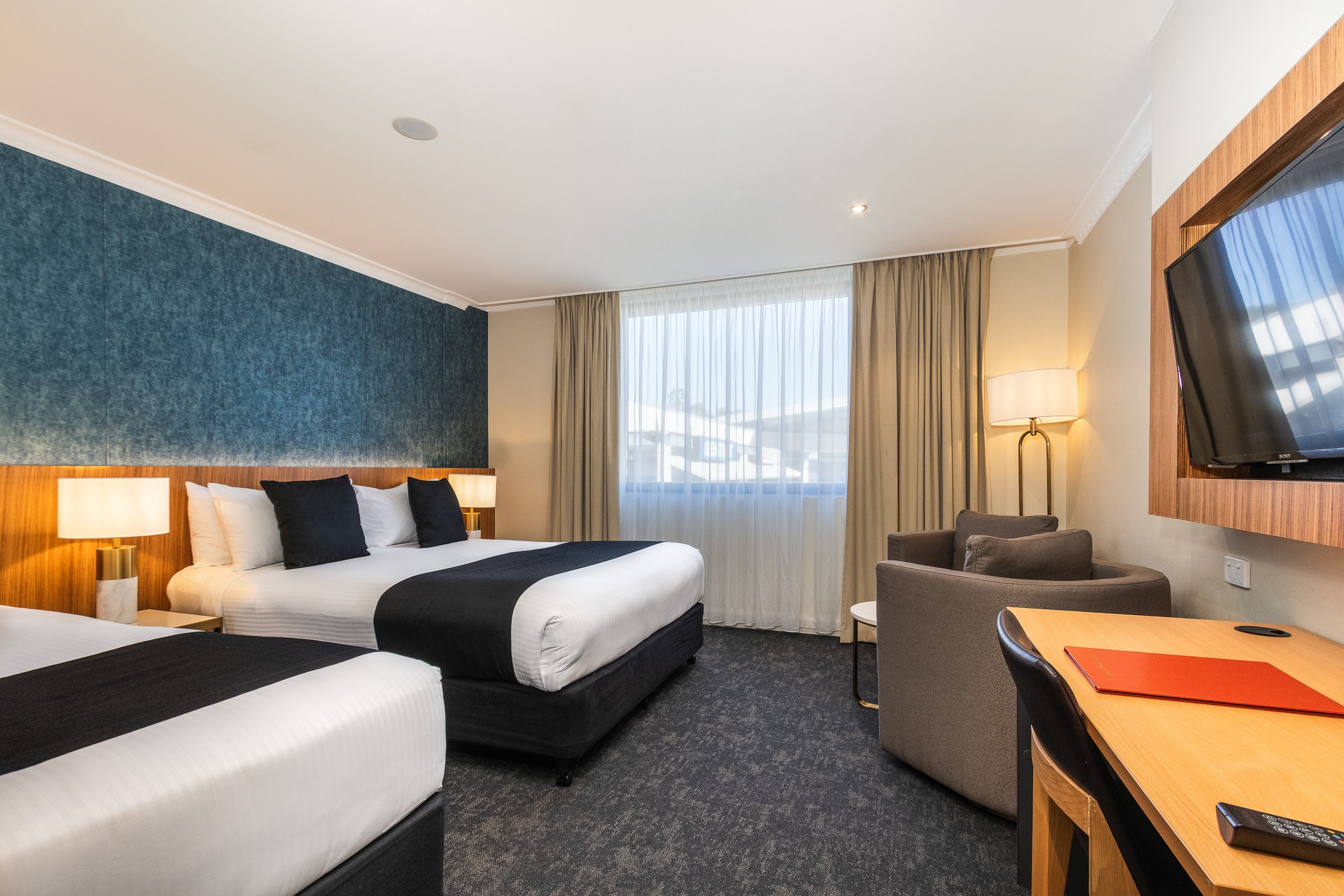 A hotel room with two beds, a window with curtains, a television mounted on the wall, a sitting area with two chairs and a small table, a desk with a closed red folder, and a lamp providing soft lighting.