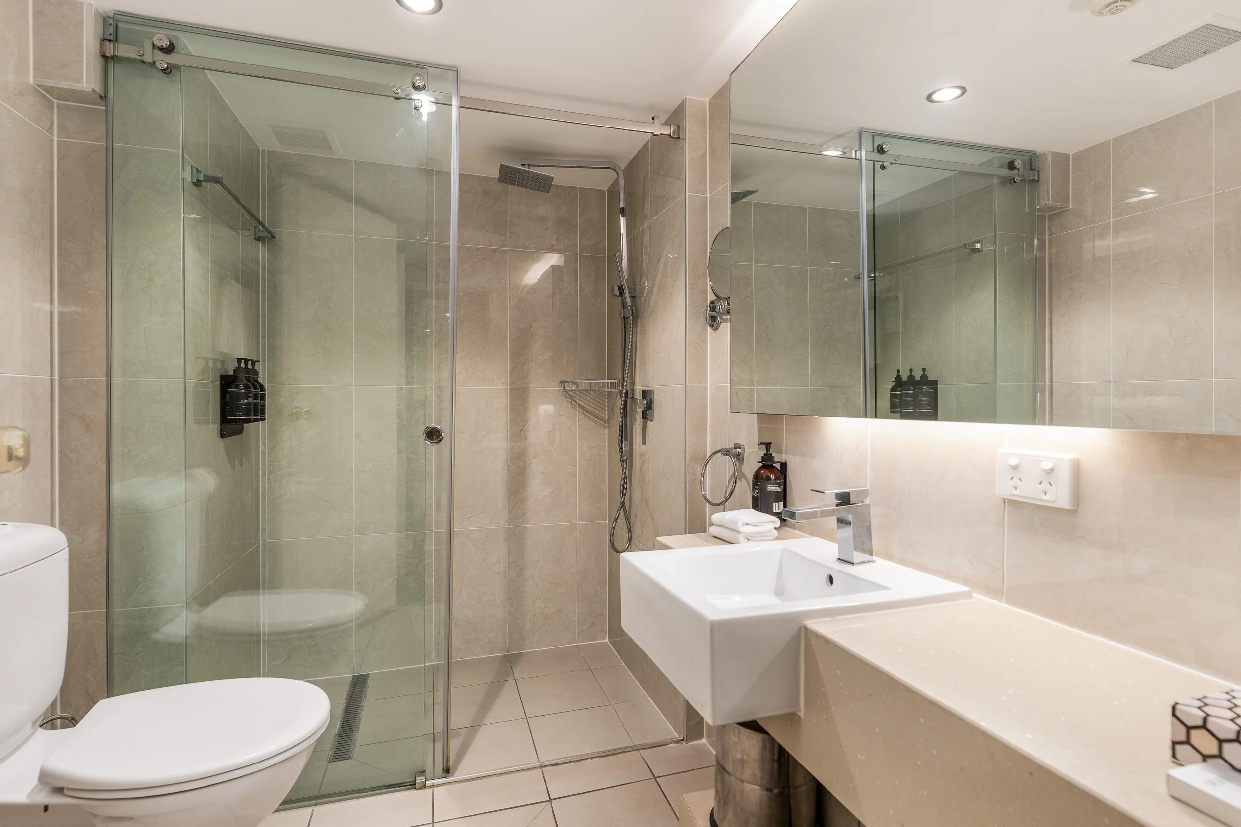 A modern bathroom with a glass shower enclosure, beige tiled walls, and a white sink with a countertop. There is a mirror above the sink, a towel, and black bottles on a small shelf inside the shower. The bathroom features recessed lighting and a wal