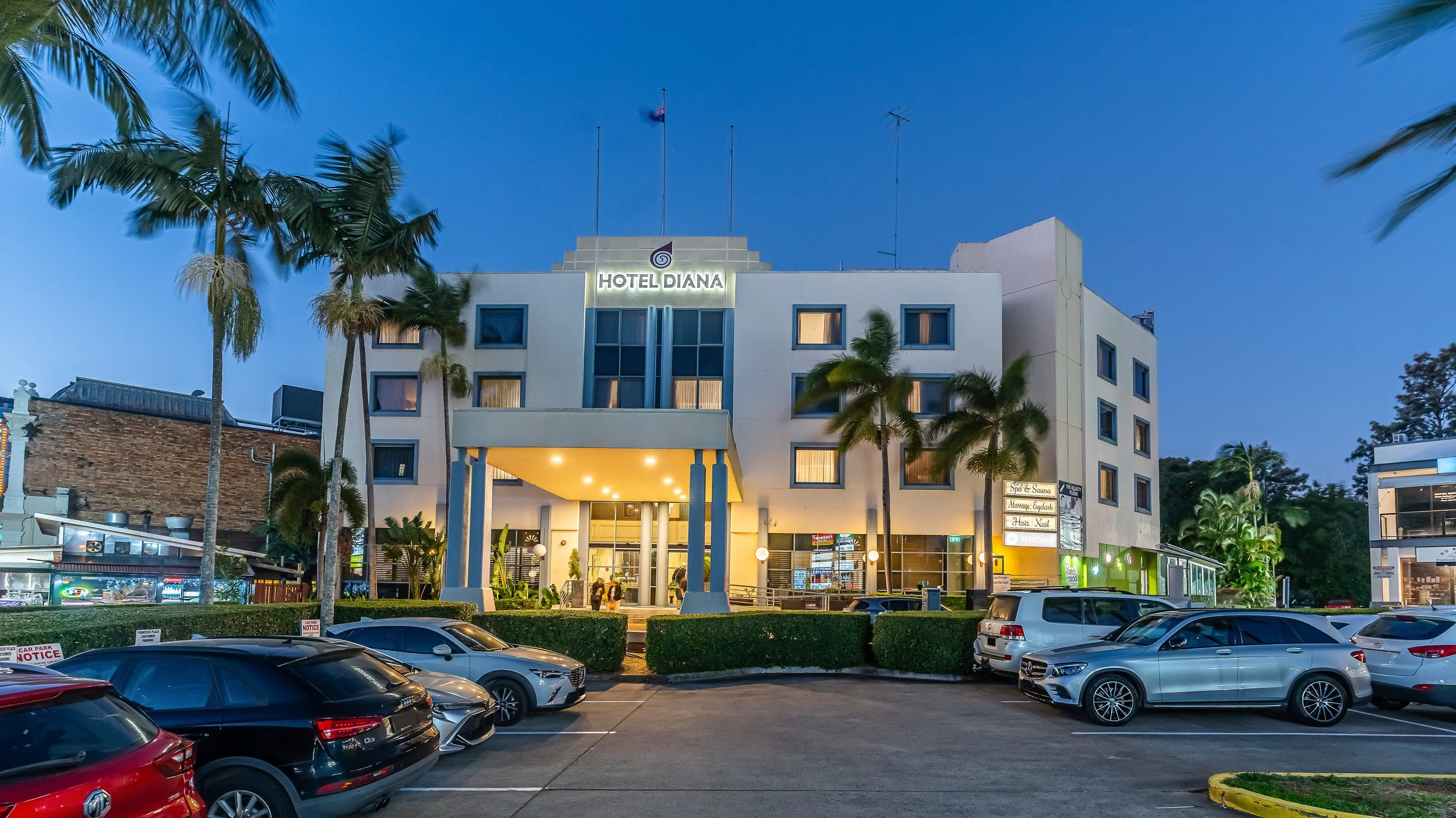 A multi-story hotel called Hotel Diana with palm trees in front, parking lot filled with cars, and a blue sky at dusk.