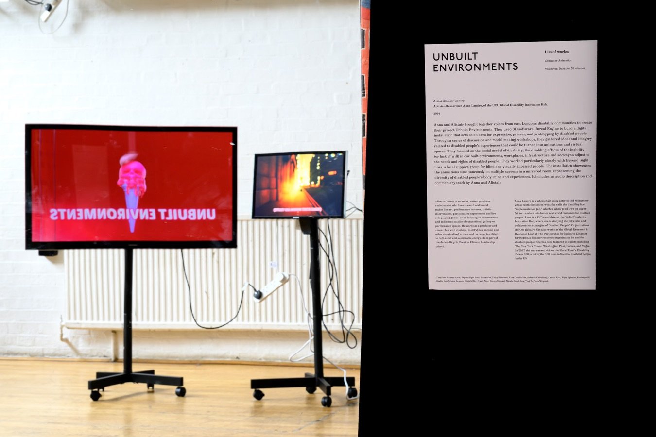 Two screens reflected in a mirror, one showing the exhibition title, Unbuilt Environments, in front of a pink ice cream cone, and the other showing an orange-lit streetscape dotted with blobs of light. Next to the mirror is an exhibition poster with