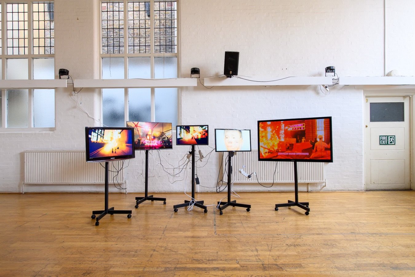 A digital art installation where animated environments built in Unreal Engine play across 5 screens. From left to right, the screens show 1) a streetscape dotted with blobs of light, 2) an uncanny, theme-park-like environment with bright pink lights,