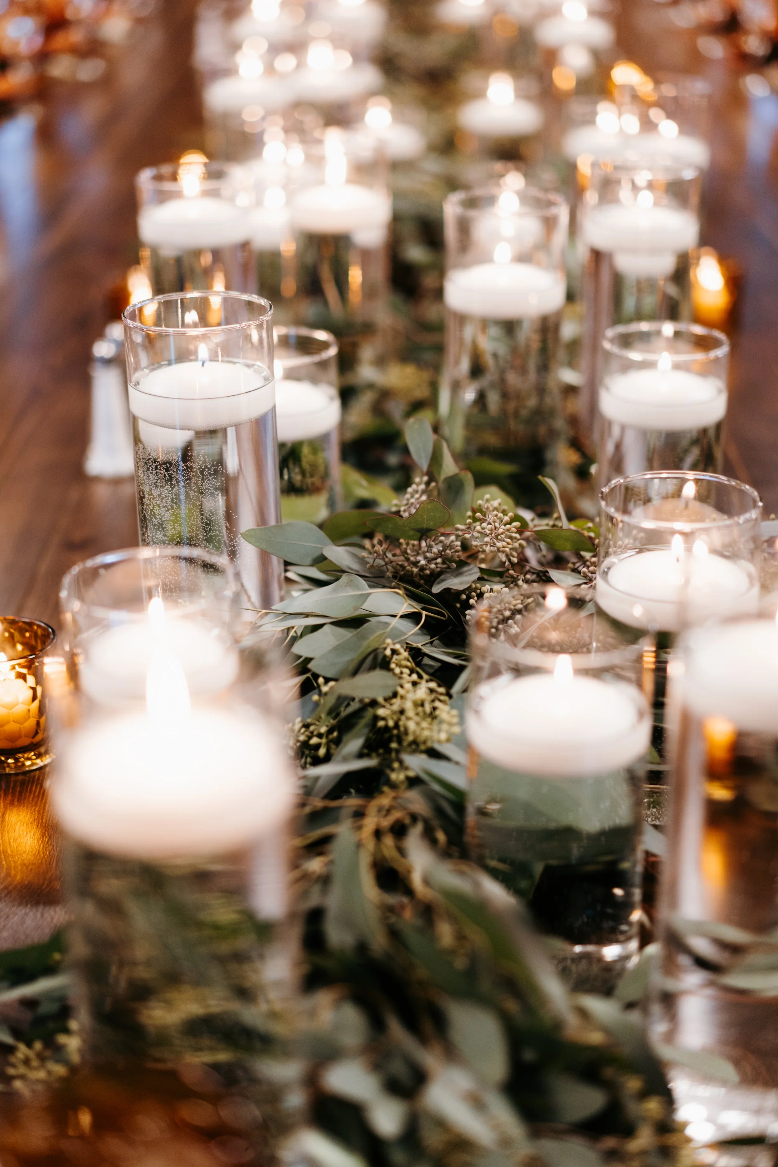 Wedding party table decorated with white candles in glass holders and surrounded by green foliage and small flowers.