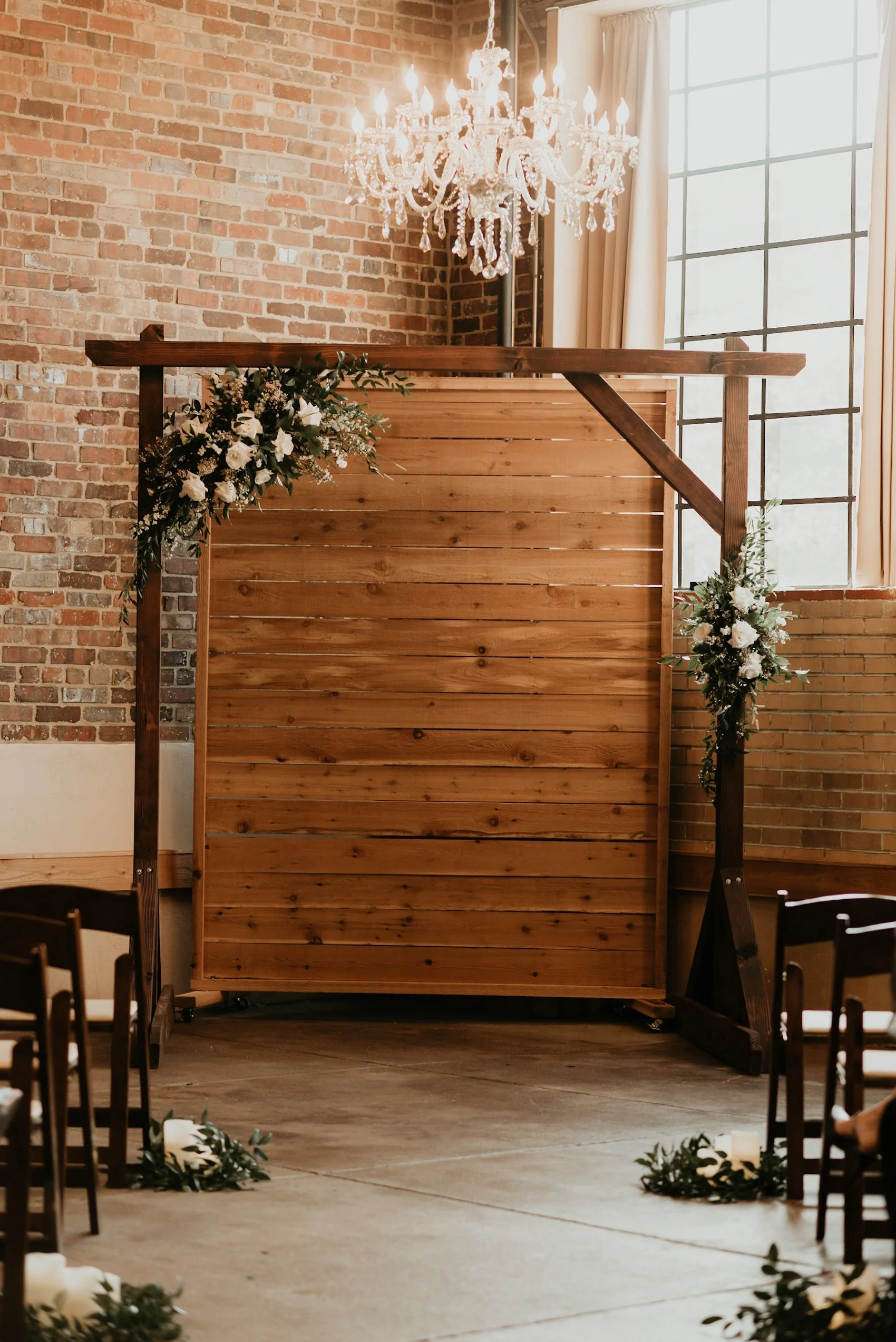Wedding ceremony backdrop at Upper Larimer wedding venue in Denver decorated with white flowers and greenery, set against a brick wall and large window, illuminated by a crystal chandelier.