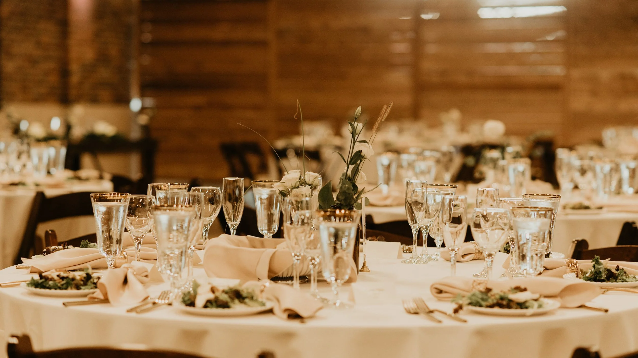 Wedding reception table decorated with glassware, floral centerpieces, and beige napkins in a rustic wooden venue.