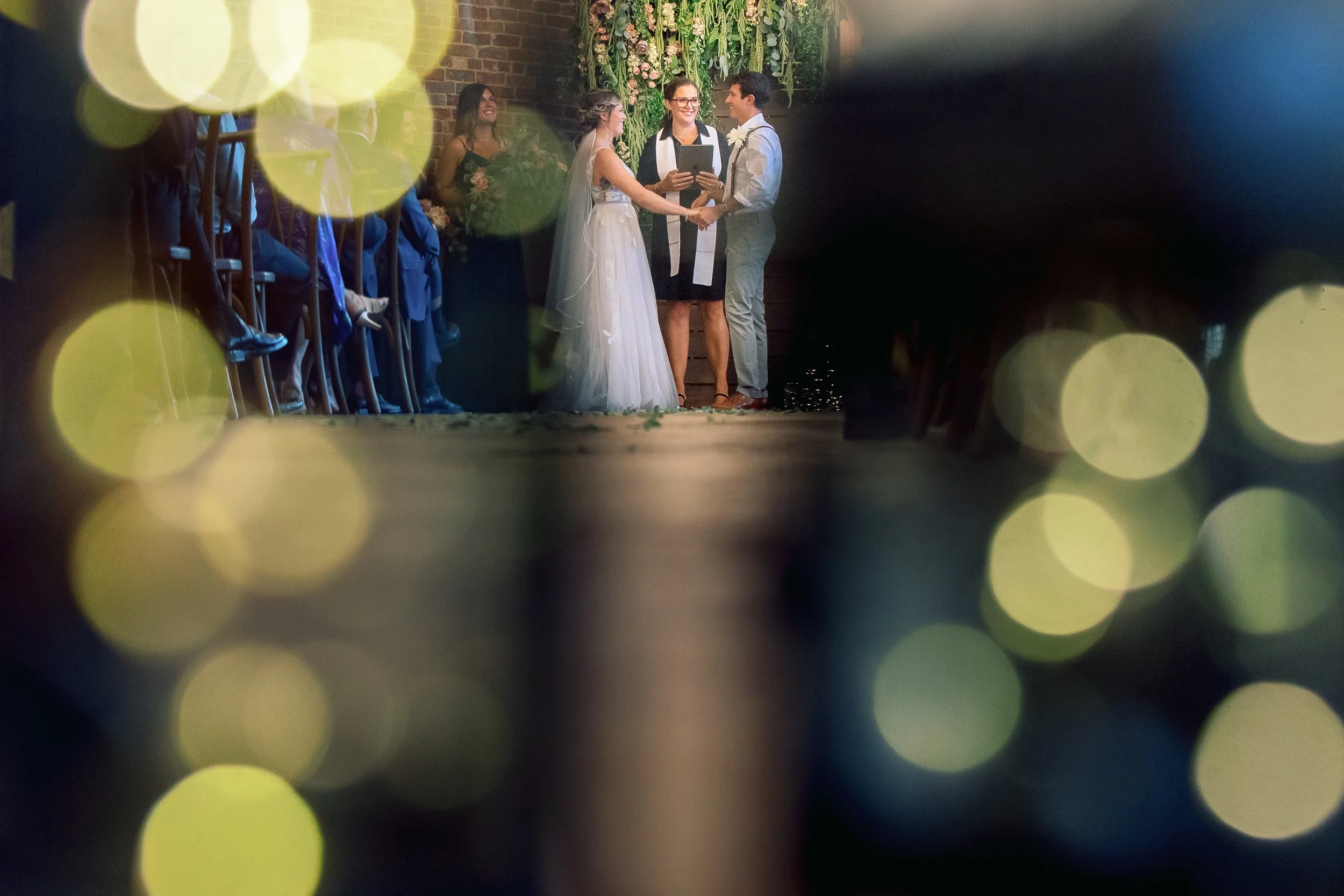 A wedding ceremony with a bride and groom exchanging vows, surrounded by friends and a wedding officiant, at Upper Larimer decorated with flowers and greenery.