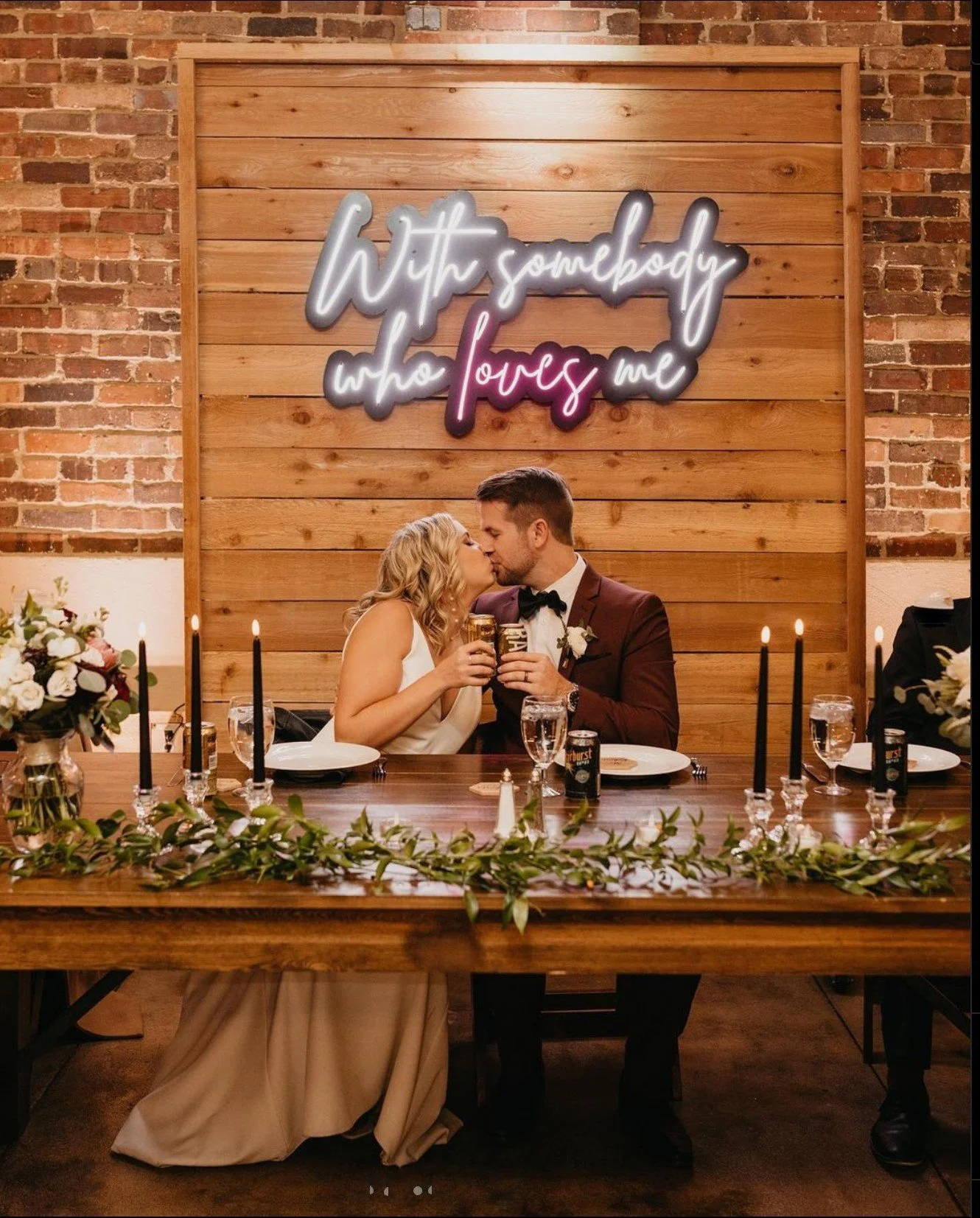 A bride and groom sitting at a wedding reception table, leaning in for a kiss while holding glasses. The background features a wooden wall with a neon sign that says 'with somebody who loves me.' The table is decorated with candles, flowers, and greenery.