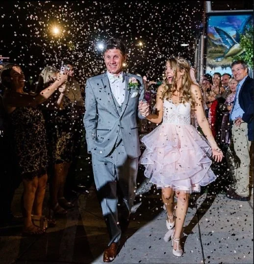 A couple walking down a party or celebration with confetti falling around, surrounded by smiling people, with colorful lights and a blue screen in the background.