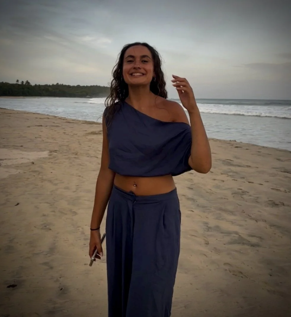 A woman with long curly hair smiling on a sandy beach near the ocean during sunset, wearing a dark blue off-shoulder top and matching wide-leg pants, holding a smartphone.