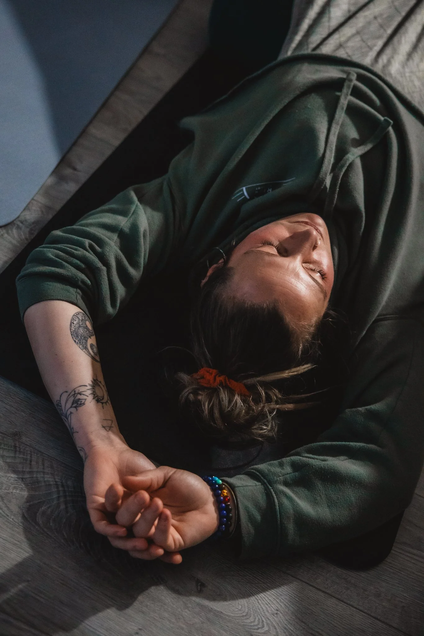 A woman with red hair practicing yoga, sitting on a yoga mat with legs crossed, holding her feet with both hands, in a room with a dark wall and wooden floor.