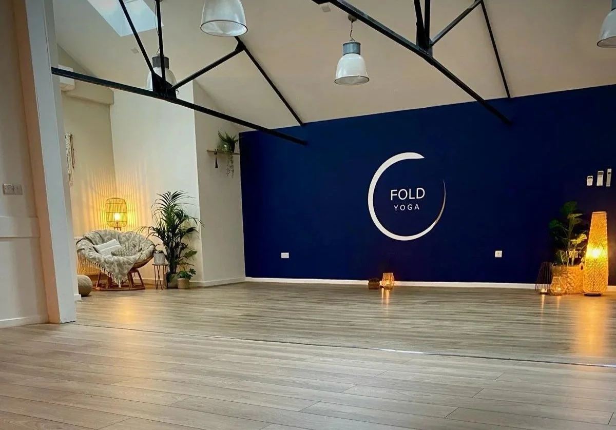 A modern Yoga Studio Macclesfield with wooden floors, navy Fold Yoga logo wall, soft lighting, and a cozy plant-filled seating area.