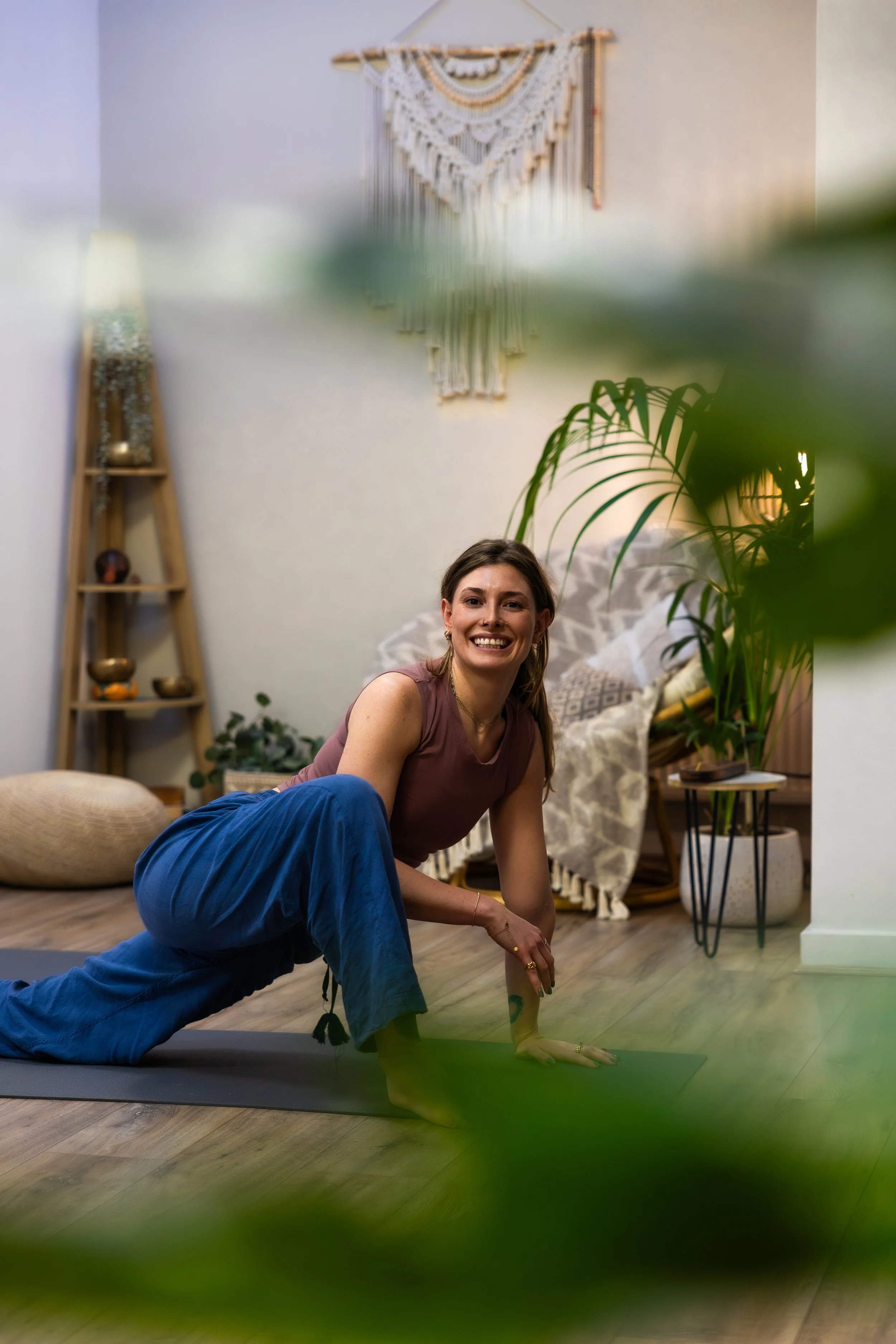 Two women sitting on the floor, laughing, with laptops and a phone in front of them. One woman is wearing a pink beanie and a gray t-shirt; the other has shoulder-length hair and a sleeveless top. A warm floor lamp and a decorative wall hanging are in the background.