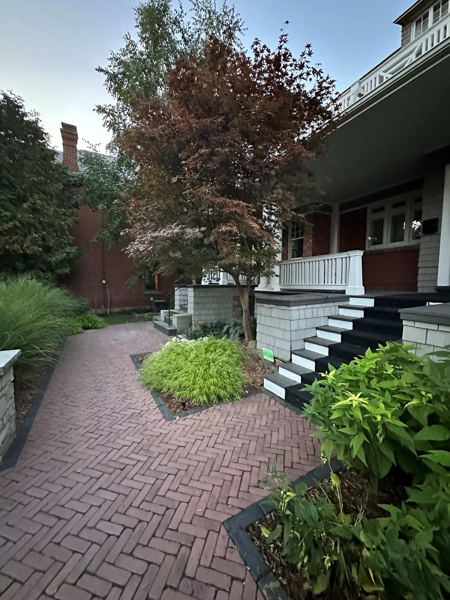 A multi-level front yard hardscape featuring a stone and brick herringbone walkway with dark stone steps, bordered by tiered natural stone retaining walls and fresh green sod.