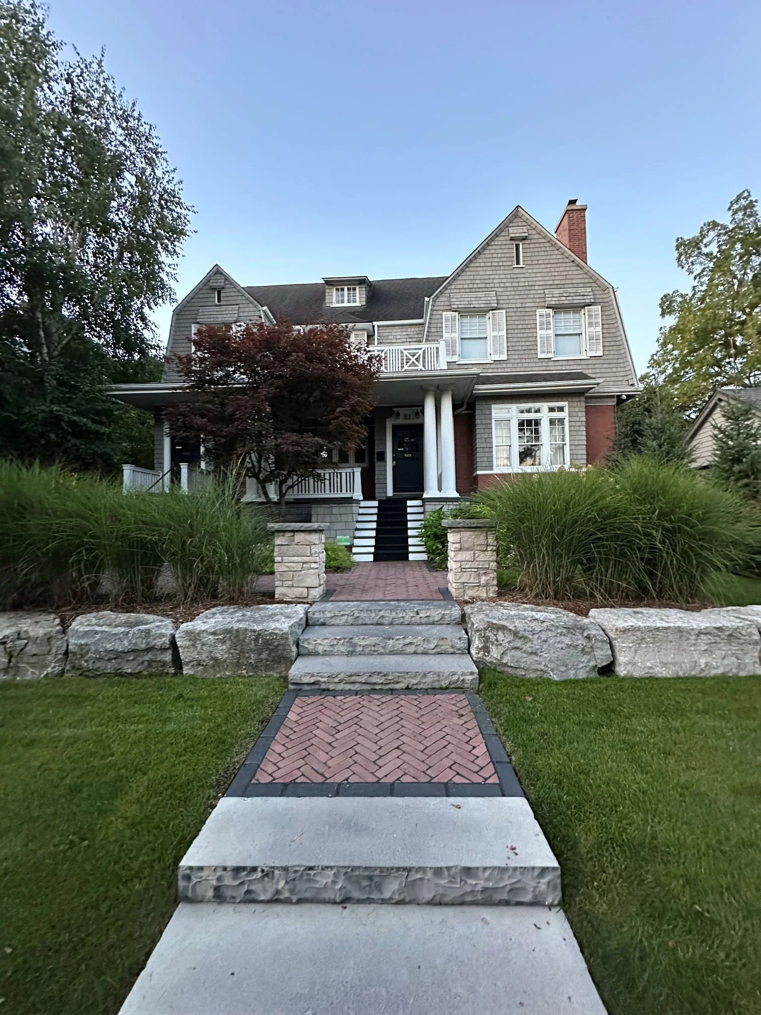 A multi-level front yard hardscape featuring a stone and brick herringbone walkway with dark stone steps, bordered by tiered natural stone retaining walls and fresh green sod.