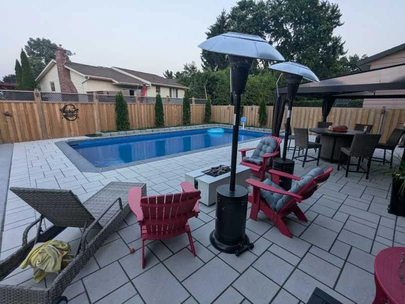 A modern backyard patio featuring Techo-Bloc Blu 60. The space includes a concrete fire pit table, and a hot tub in the background, all enclosed by a wooden privacy fence.