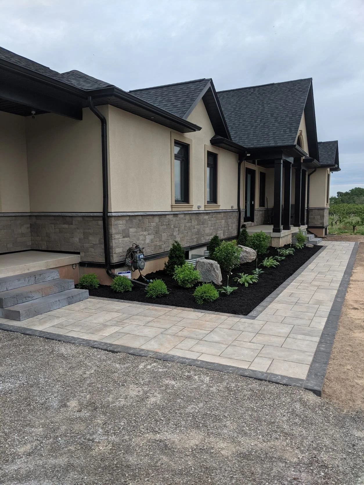 Modern residential landscape design featuring a black mulch garden bed with manicured shrubs, natural stone boulders, and a multi-tonal interlocking paver walkway 