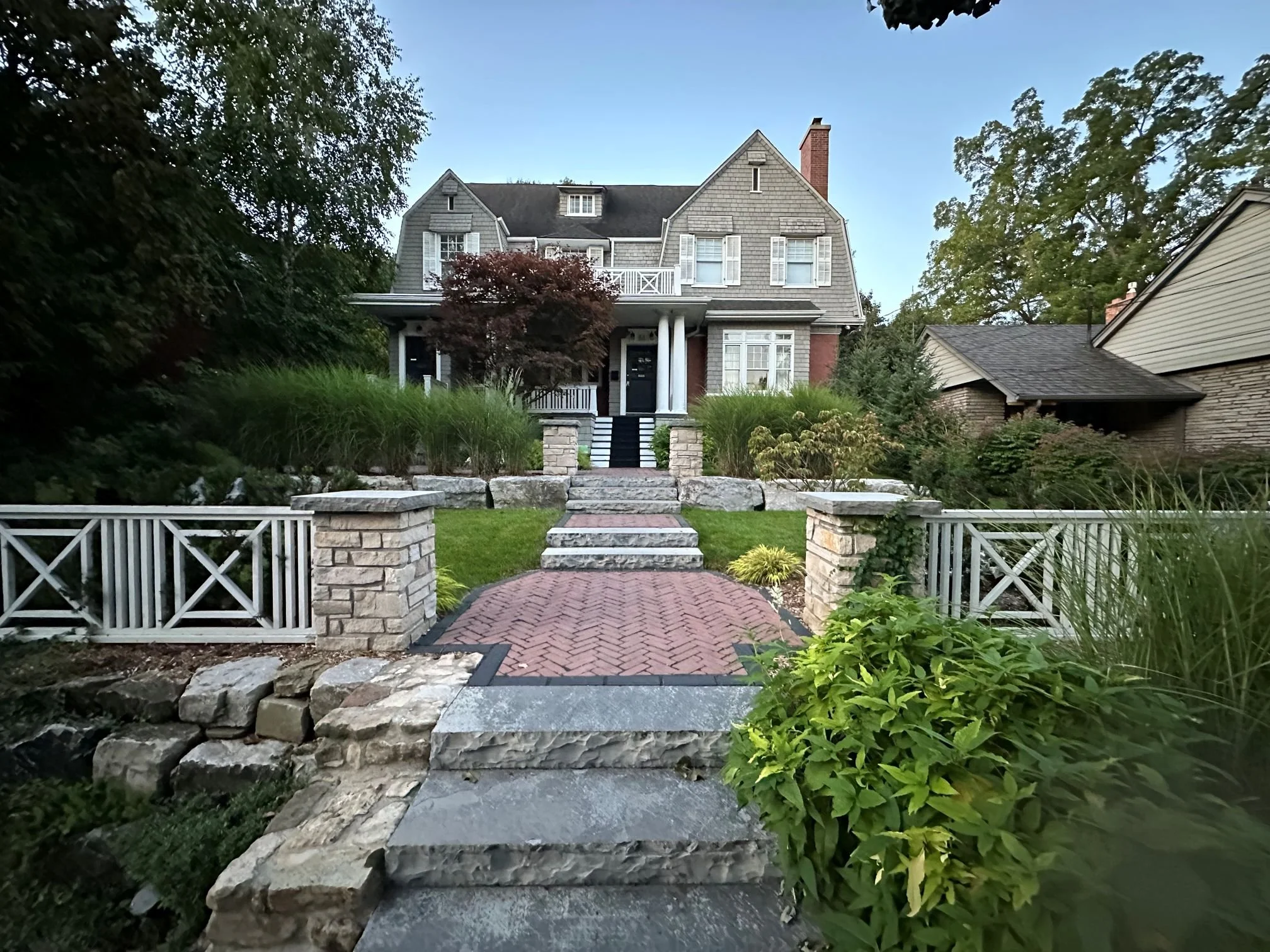 A multi-level front yard hardscape featuring a stone and brick herringbone walkway with dark stone steps, bordered by tiered natural stone retaining walls and fresh green sod.