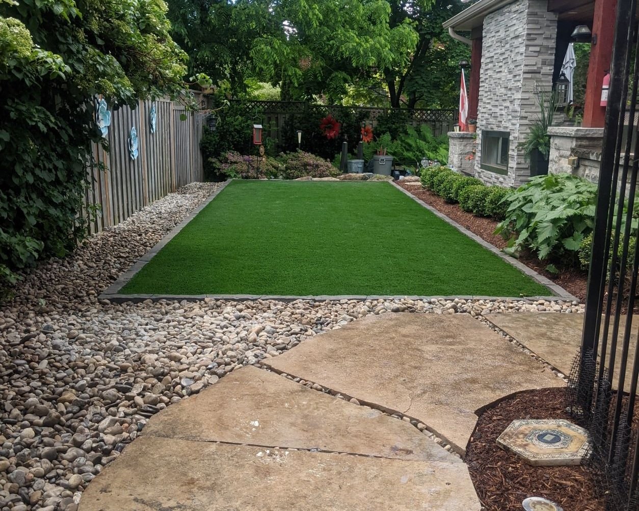 A professionally landscaped backyard featuring a rectangular lush green artificial turf lawn bordered by stone edging. The surrounding area includes light-colored river rock, mulch, and large natural flagstone pavers.