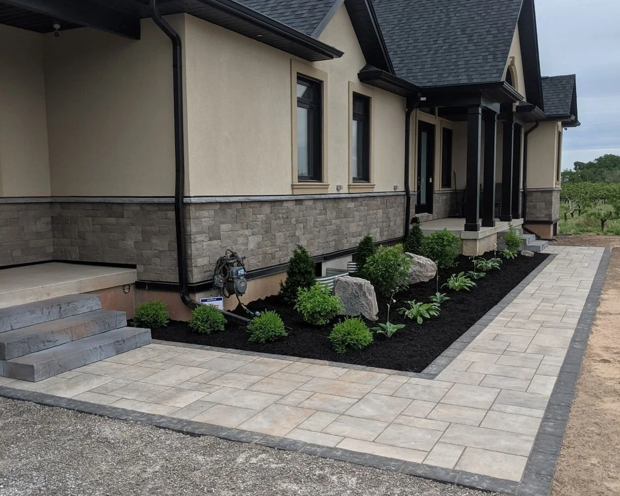 Modern residential landscape design featuring a black mulch garden bed with manicured shrubs, natural stone boulders, and a multi-tonal interlocking paver walkway 