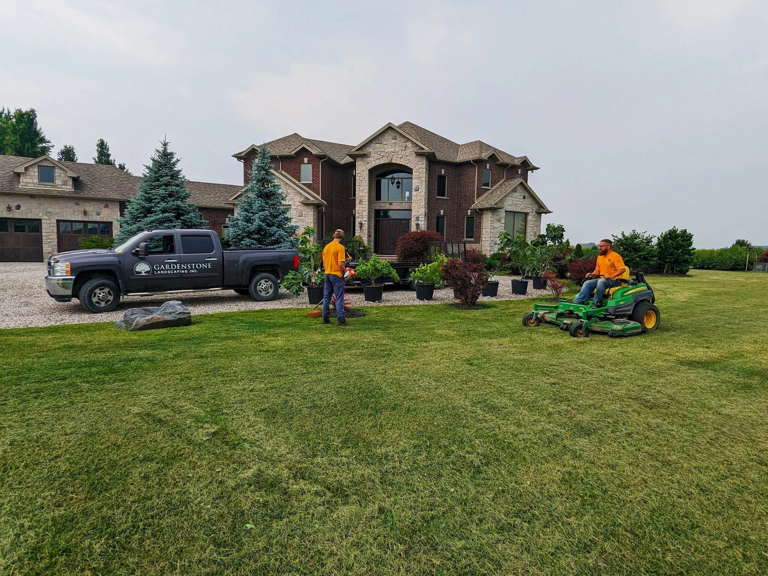 Two professional landscapers from Gardenstone Landscaping Inc. performing lawn maintenance and garden installation at a large residential estate.