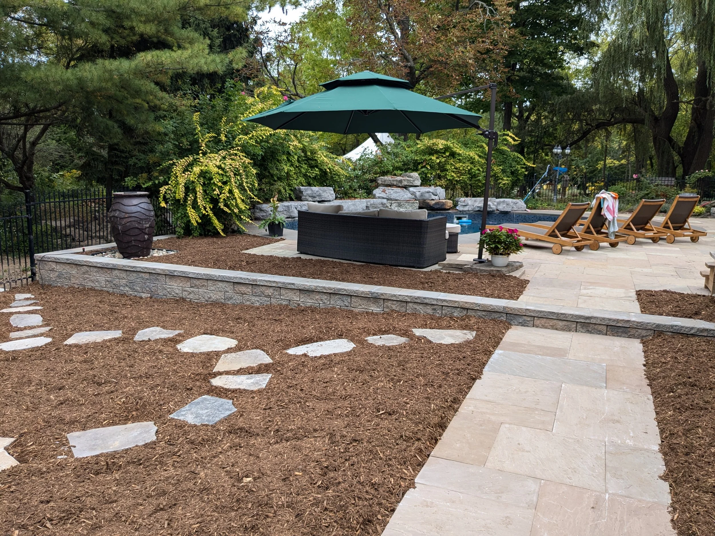 Professional backyard landscaping design including custom stonework, a stone retaining wall, flagstone walkways, and a luxury pool deck with outdoor furniture in Grimsby.
