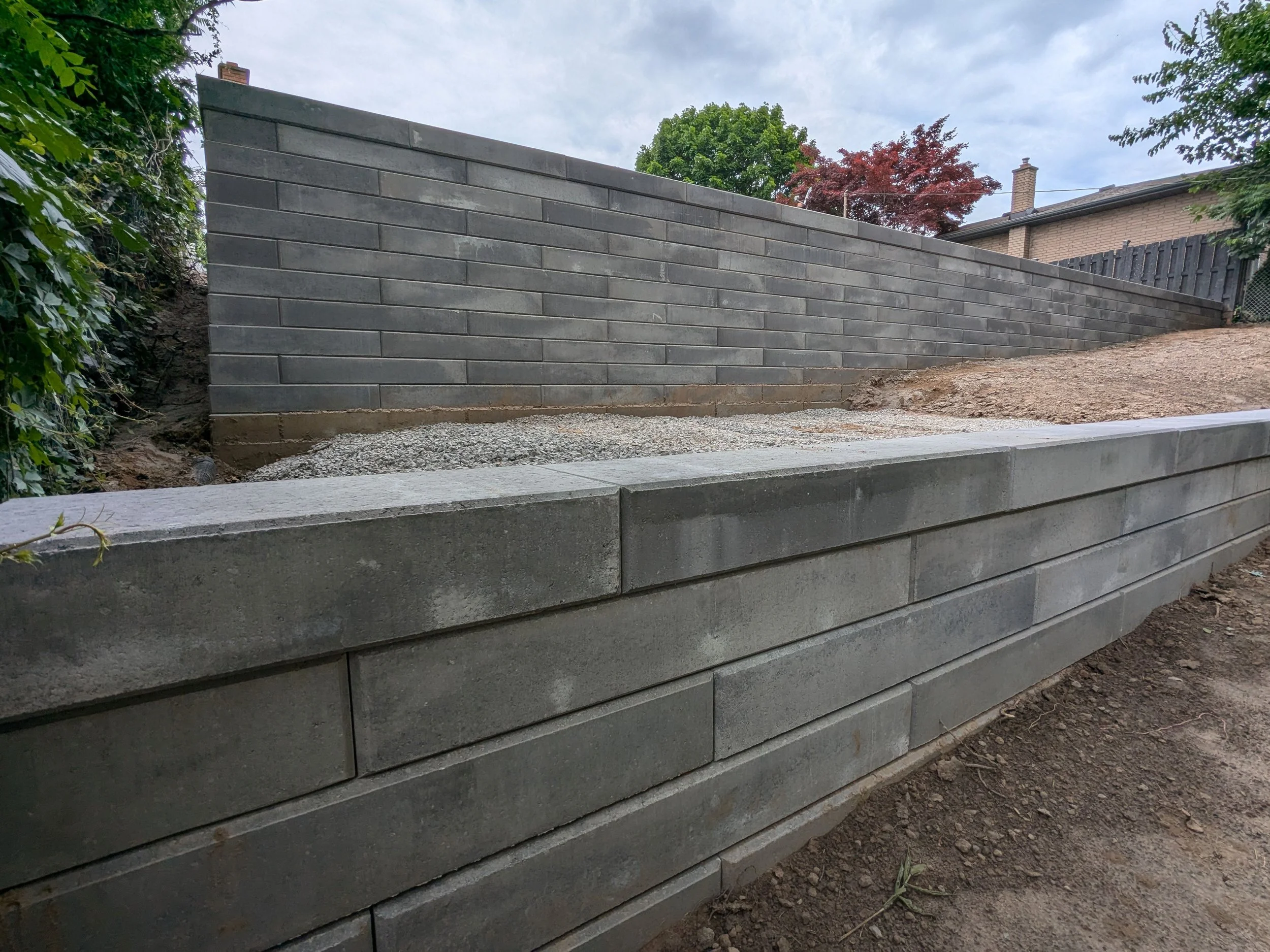 Modern tiered retaining wall installation using Unilock Siena stone blocks. The project features a large upper wall and a smaller lower tier, showcasing a clean and structural backyard landscape design in St. Catharines.