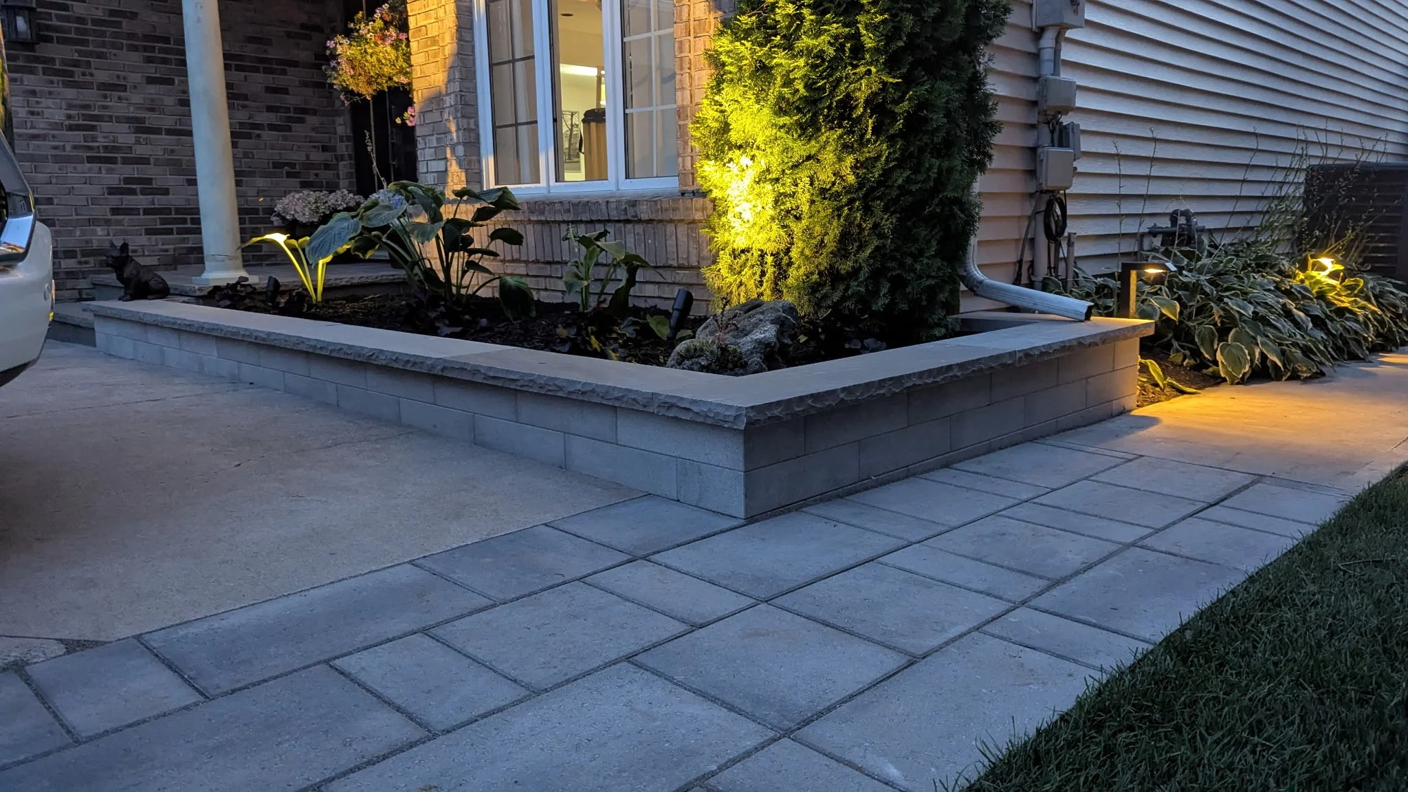 A modern stone retaining wall and tiered garden bed featuring integrated landscape lighting, a gray paver walkway, and lush greenery.
