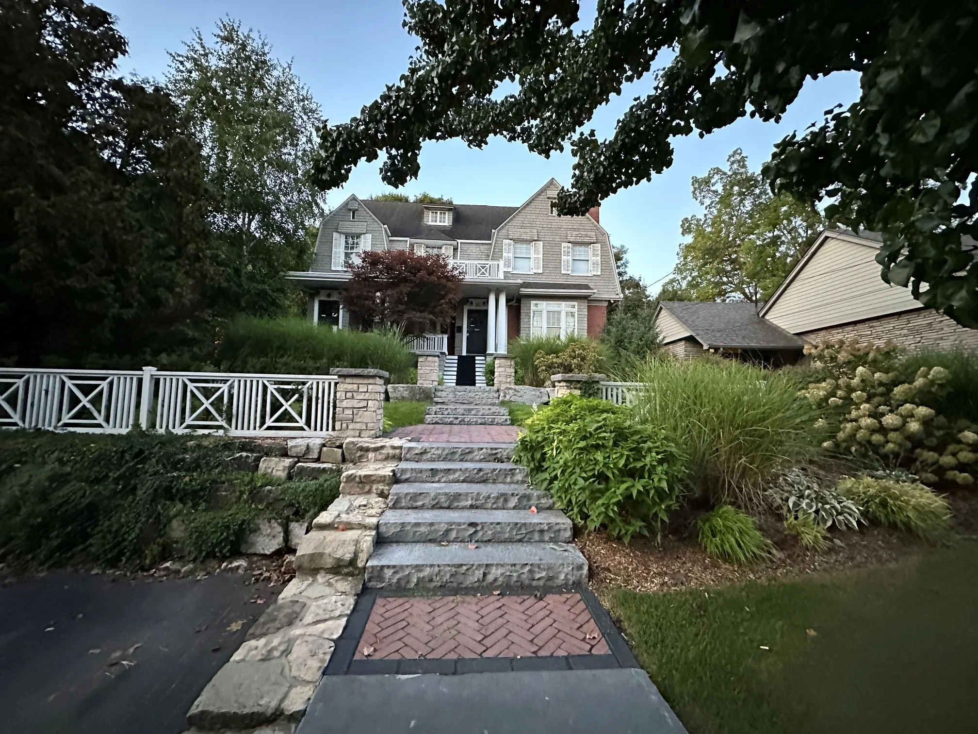 A multi-level front yard hardscape featuring a stone and brick herringbone walkway with dark stone steps, bordered by tiered natural stone retaining walls and fresh green sod.