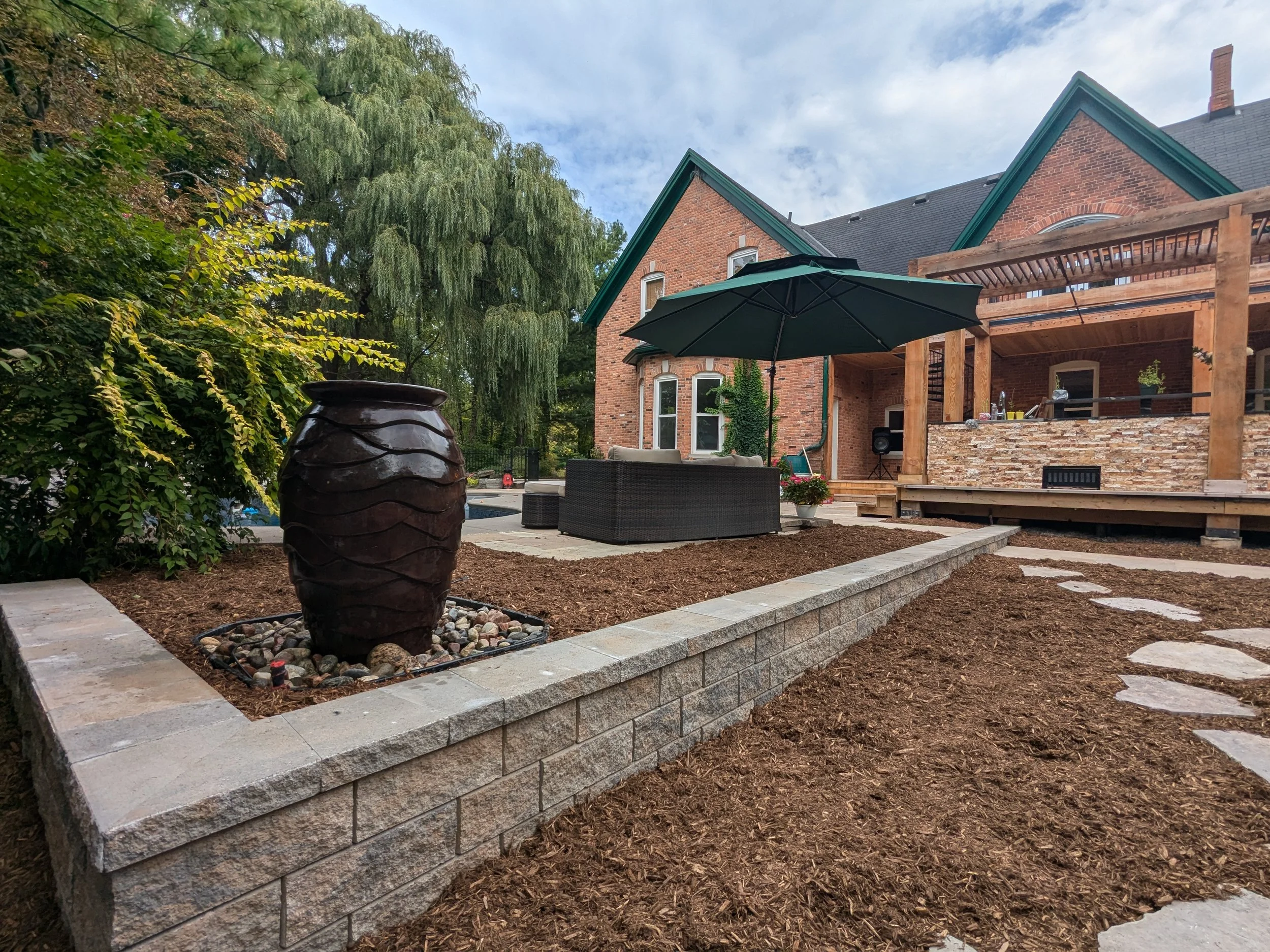 A professionally landscaped backyard featuring a tiered garden bed with walls made from Techo-Bloc Semma wall block, dark wood mulch, and a large decorative ceramic water urn.