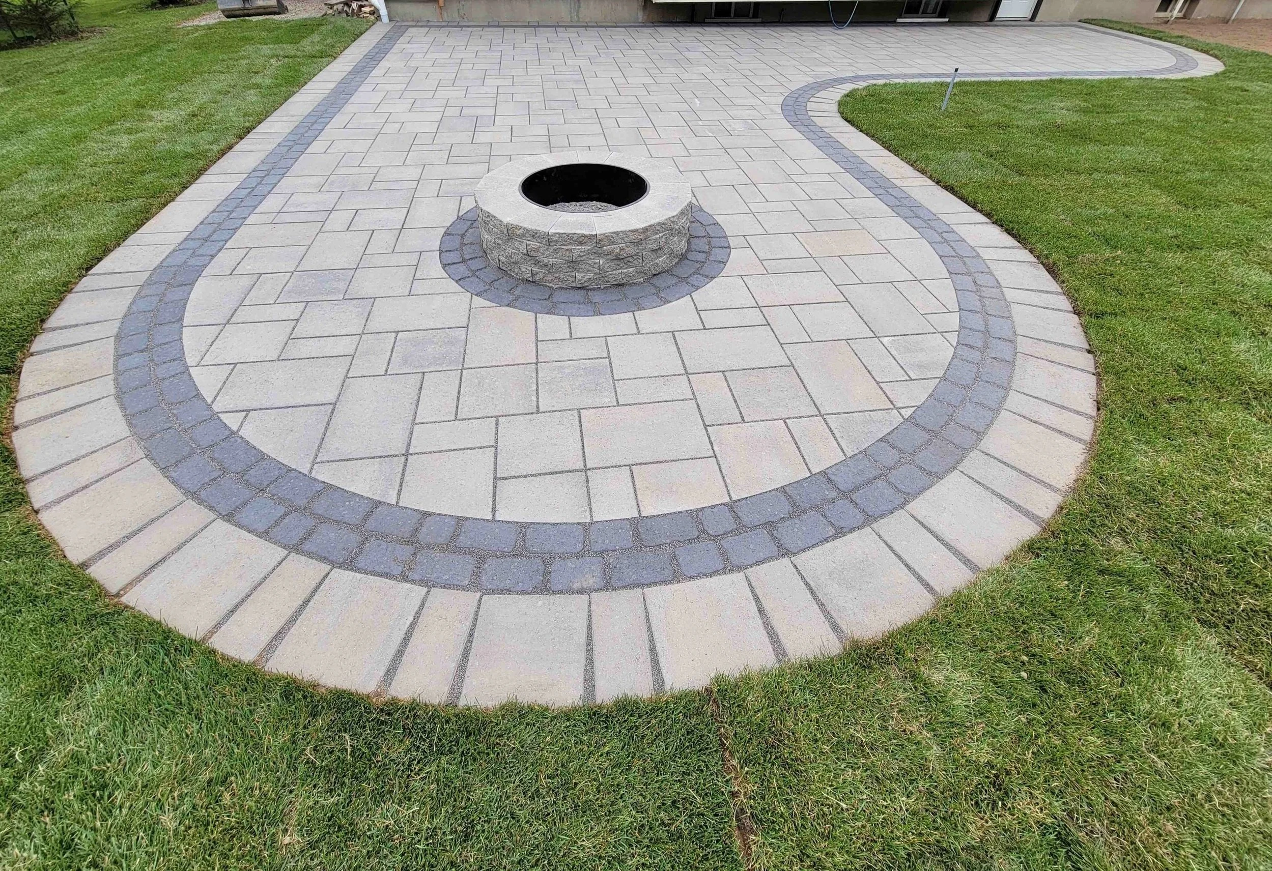 A curved backyard patio featuring Techo-Bloc Blu 60 pavers in Champlain Grey, accented by a onyx black Villagio cobble border and a central stone fire pit