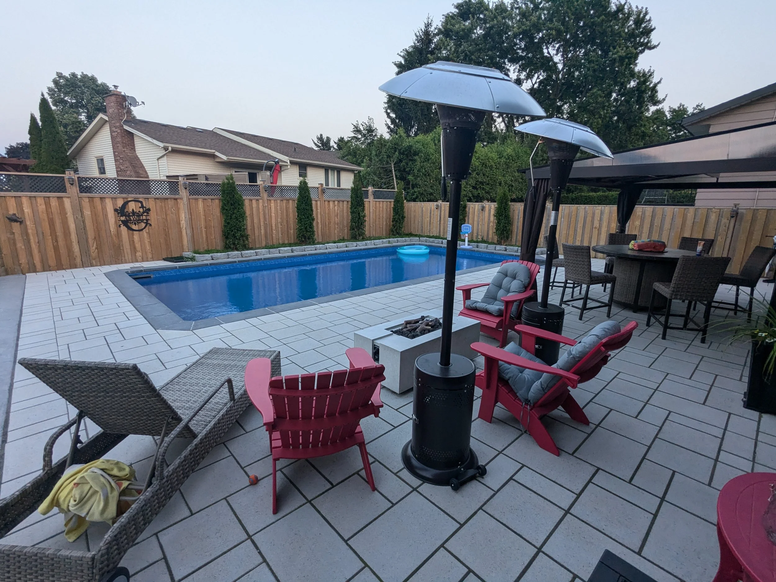 A modern backyard patio featuring Techo-Bloc Blu 60. The space includes a concrete fire pit table, and a hot tub in the background, all enclosed by a wooden privacy fence.