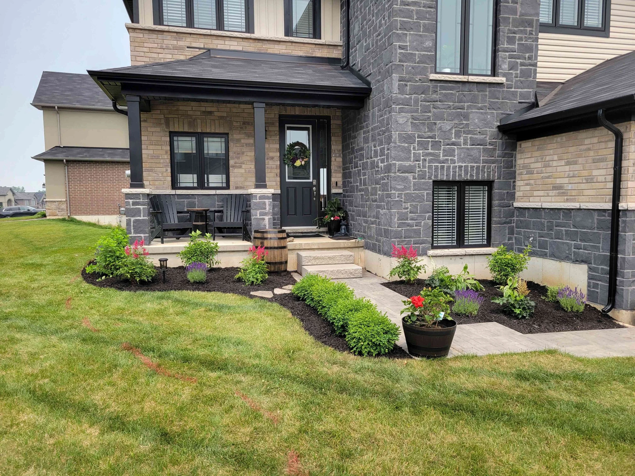 A modern home exterior featuring a well-maintained garden bed with dark mulch, pink astilbe, lavender, and a row of manicured green shrubs bordering a stone walkway.